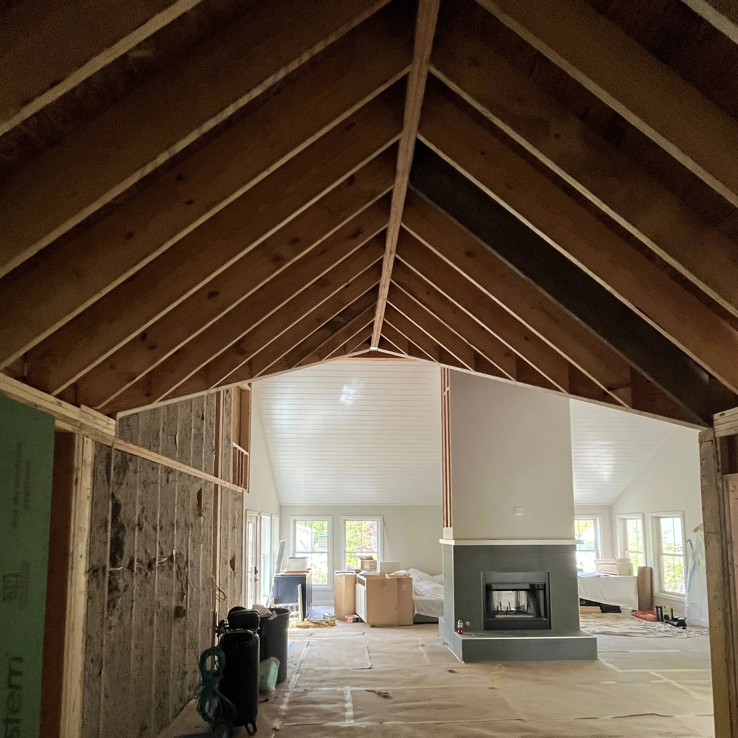 Interior view of a house under construction, showing exposed wooden framing, a partially finished ceiling, and large windows in the background letting in natural light.