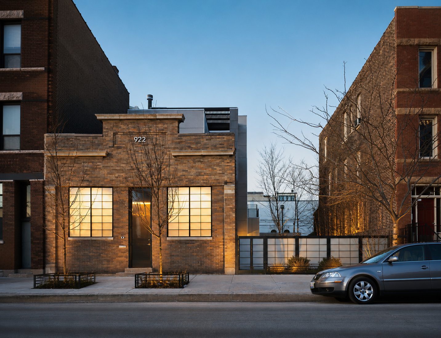 A modern brick building with large lit windows and the number 922 on the facade. The building is flanked by leafless trees, with a sidewalk and parked car in the foreground. The sky is clear and there are neighboring buildings.