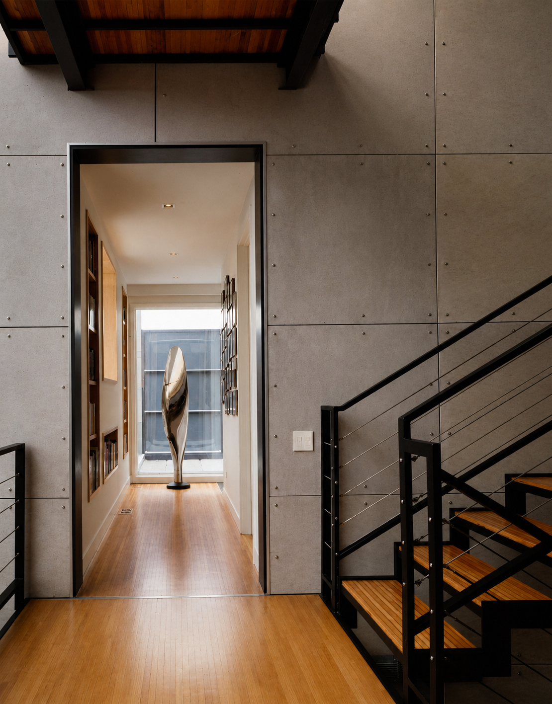 Modern interior space featuring a staircase with black metal railing and wooden steps, beige textured wall with exposed screws, and a hallway with built-in bookshelf and a metallic sculpture at the end near a large window.