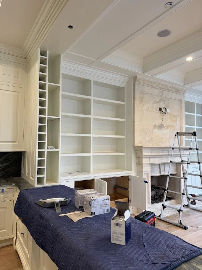 Living room with built-in white bookshelves, a stone fireplace, an adjustable ladder, and a table covered with a dark blue quilt, holding boxes and tools for home renovation.