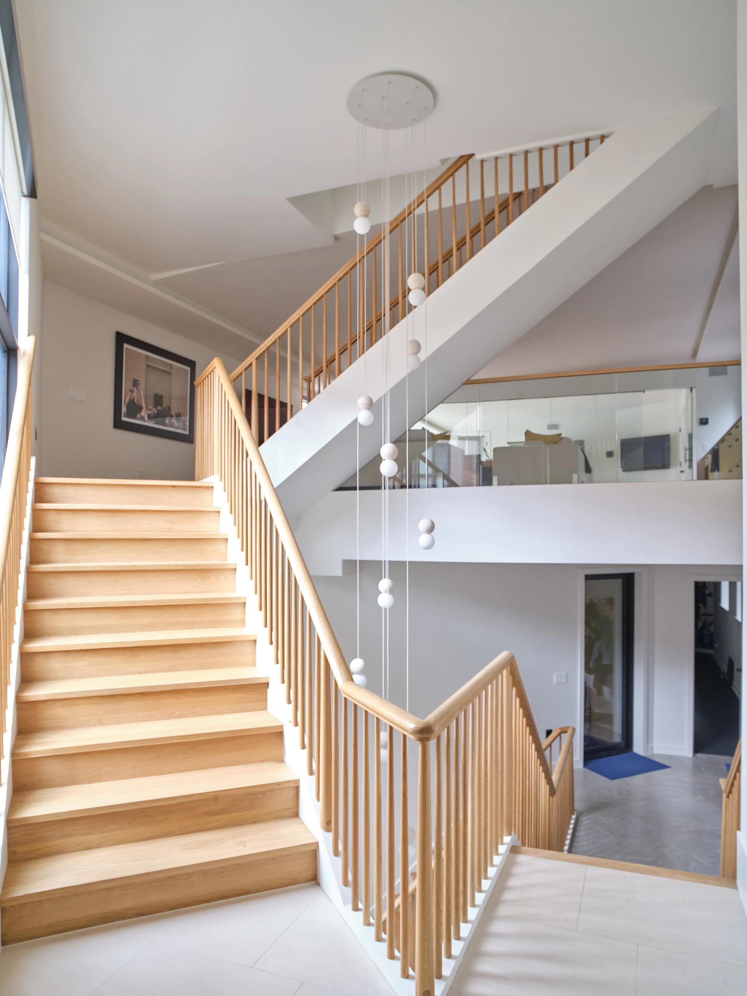 Interior view of a modern staircase with wooden steps and railing, a hanging light fixture with round bulbs, and a glass wall showing rooms inside the house.