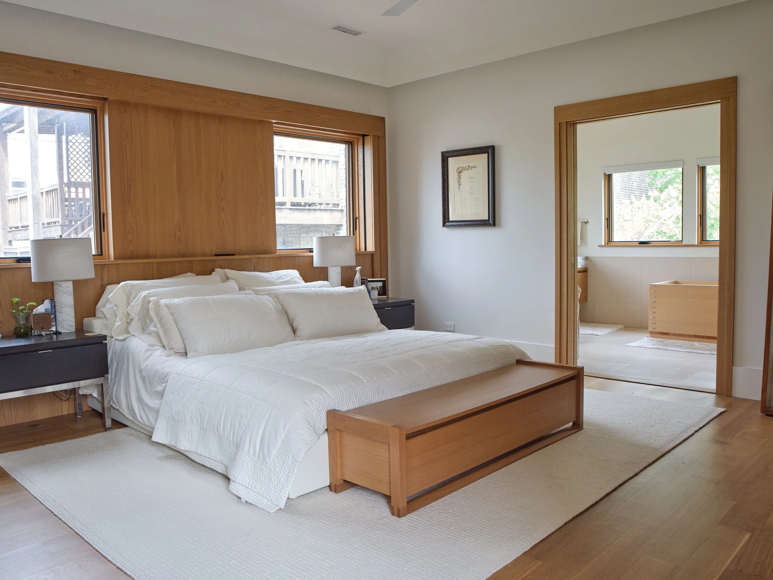 A bedroom with a bed, two nightstands with lamps, and framed artwork on the wall, featuring two windows with wooden trim and a door leading to an adjacent room with a smaller window.