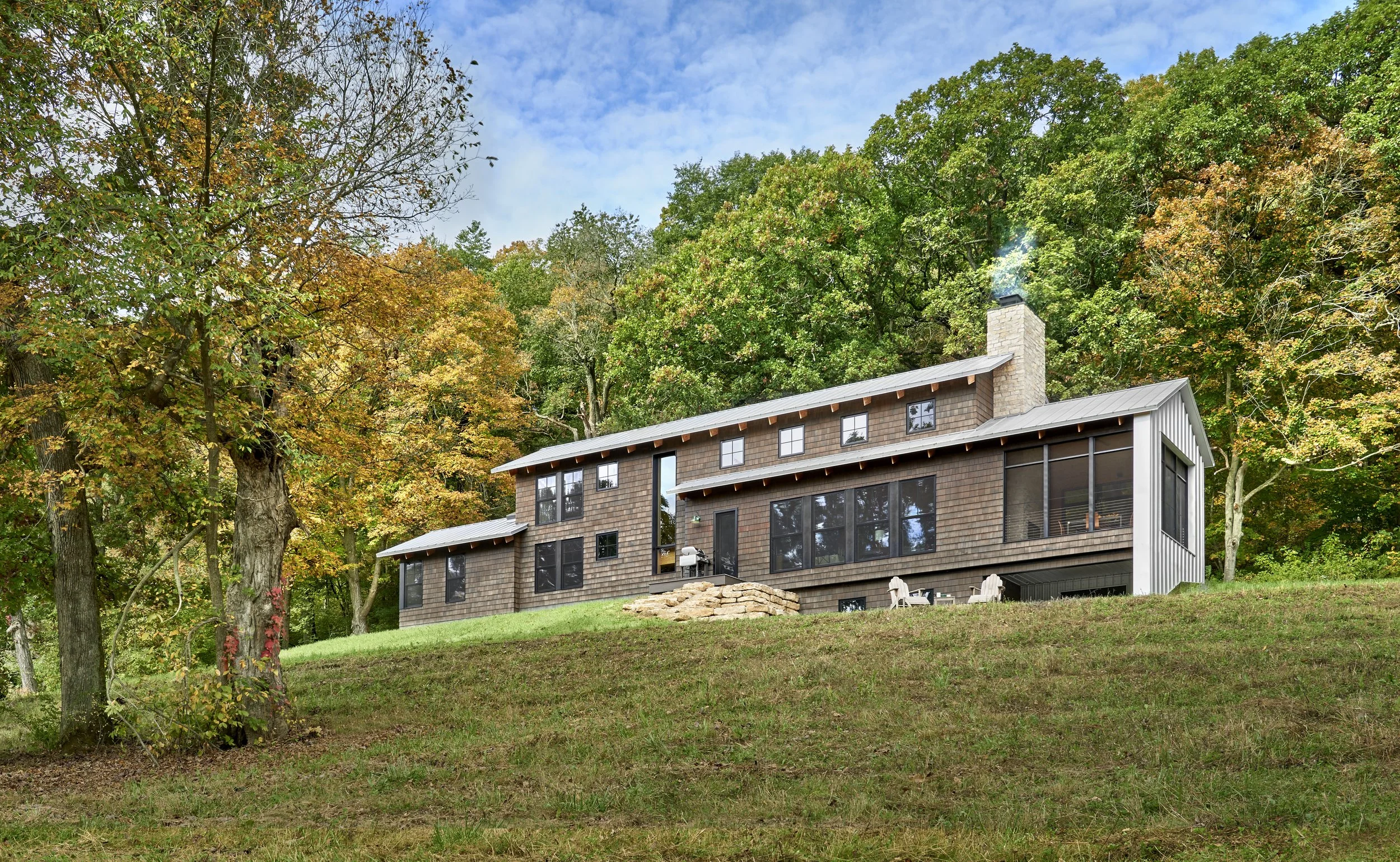 View of a house with black exterior walls, a metal roof, and large windows, surrounded by trees with autumn-colored leaves, under a partly cloudy sky.
