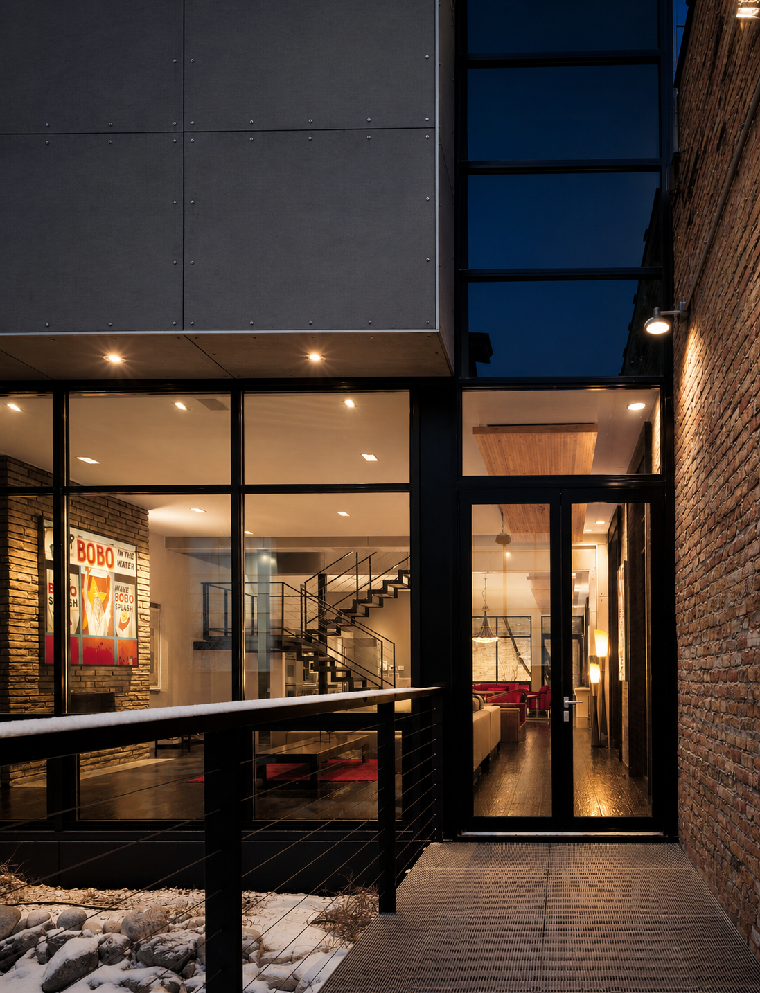 Exterior view of a modern building at night, showing large glass windows revealing a stylish interior with a staircase, seating area, and artwork, with warm lighting and brick walls.