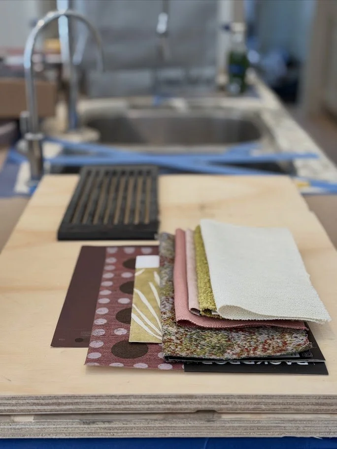 Color samples and fabric swatches on a wooden table, with a kitchen sink and faucet in the background.