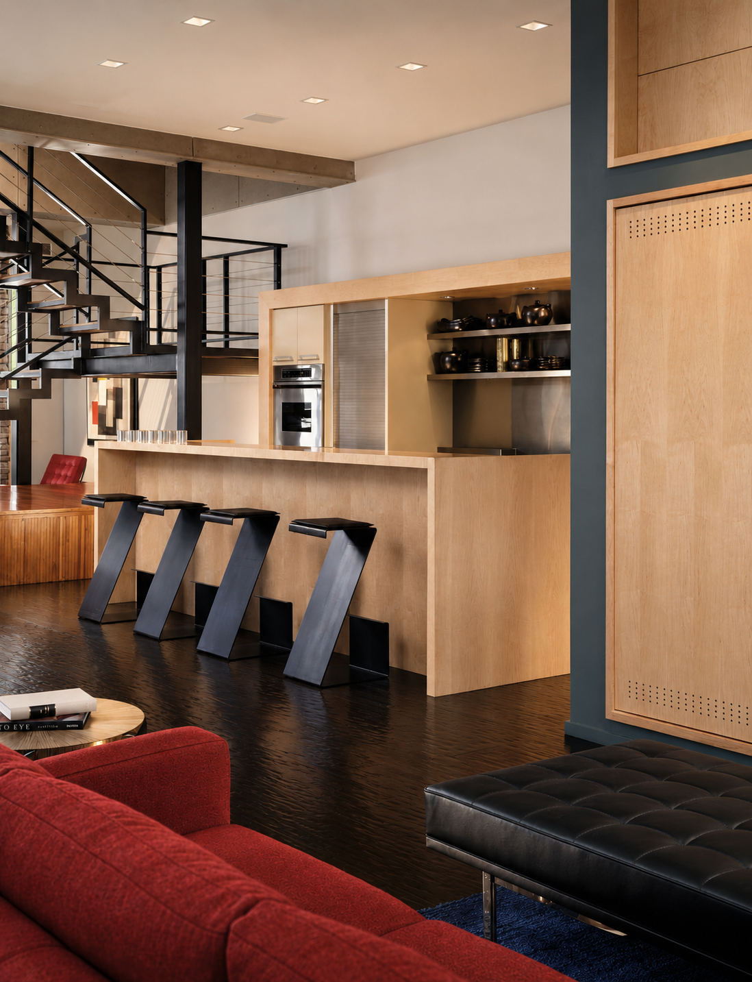 Modern kitchen with light wood cabinets, open shelves, black pottery, stainless steel oven, breakfast bar with black stools, view of staircase, and part of living room with red sofa and black ottoman.