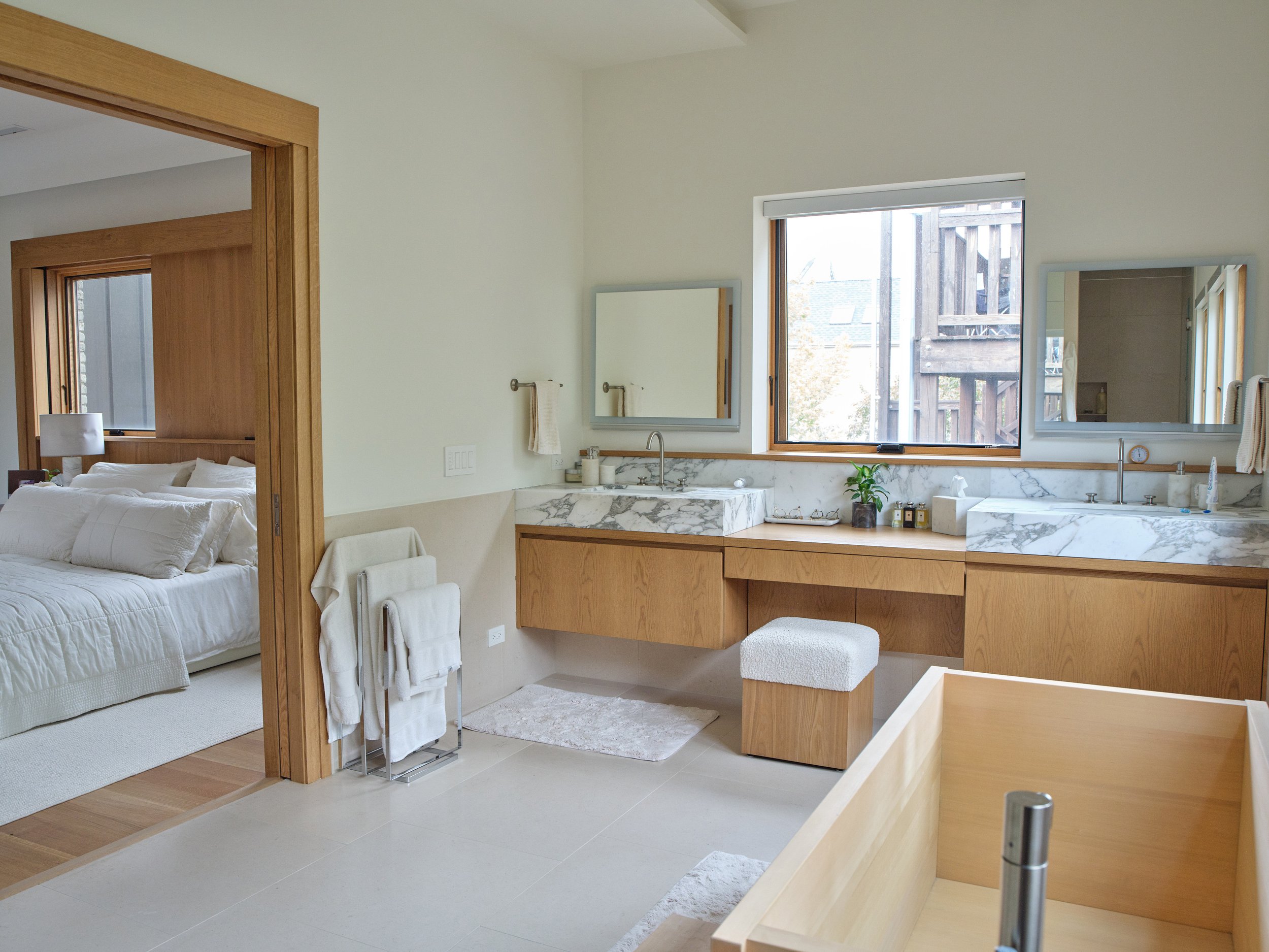 A modern bathroom with a large window overlooking a wooden balcony, marble sink, mirror, and white towels. Adjacent to the bathroom, there is a bedroom with a white bed, bedside table, and lamps.