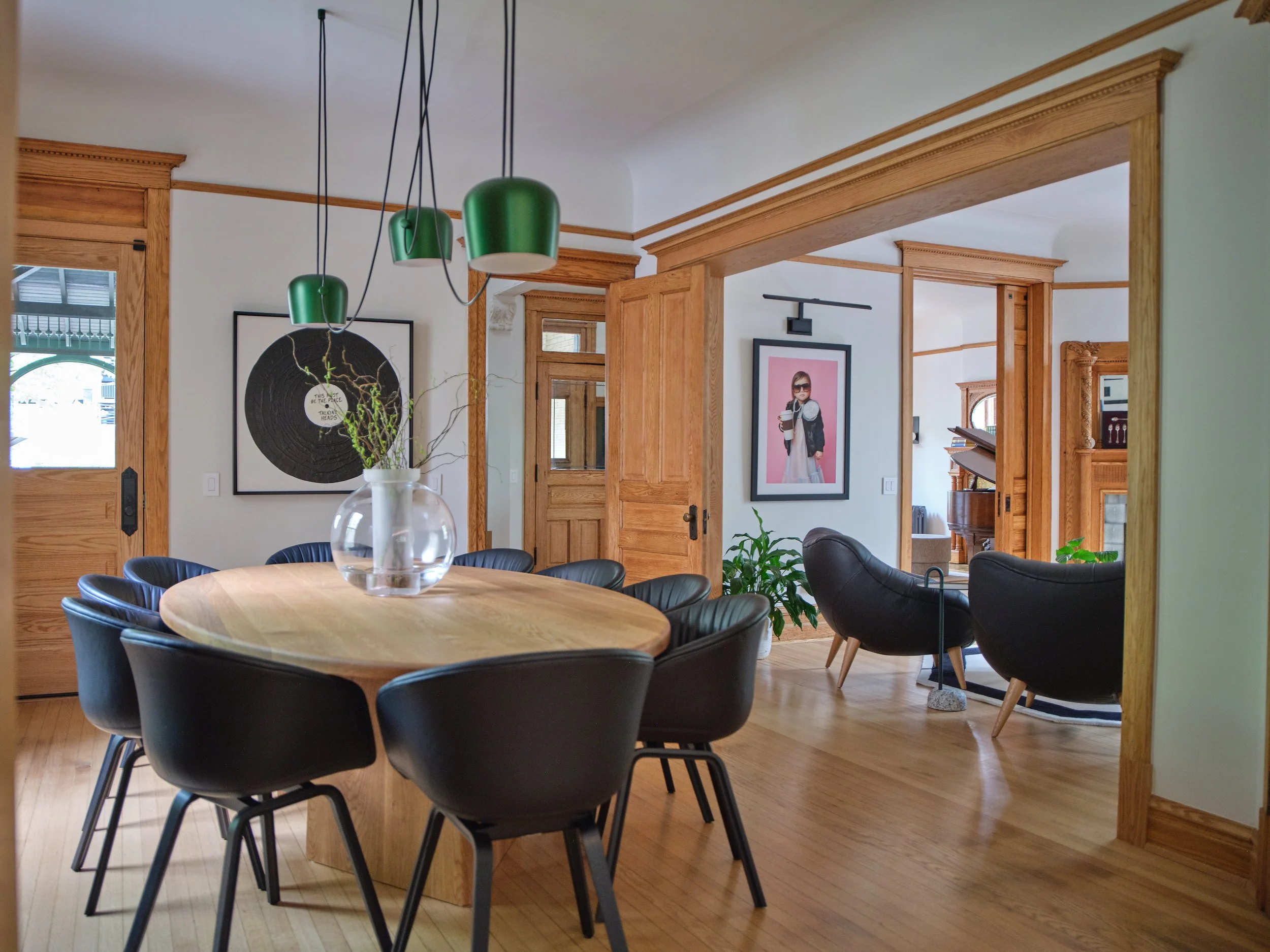 Interior view of a dining room with a round wooden table surrounded by black chairs. The room features wooden trim and door frames, with artwork on the walls, including a piece with a woman wearing sunglasses. Green pendant lights hang from the ceili