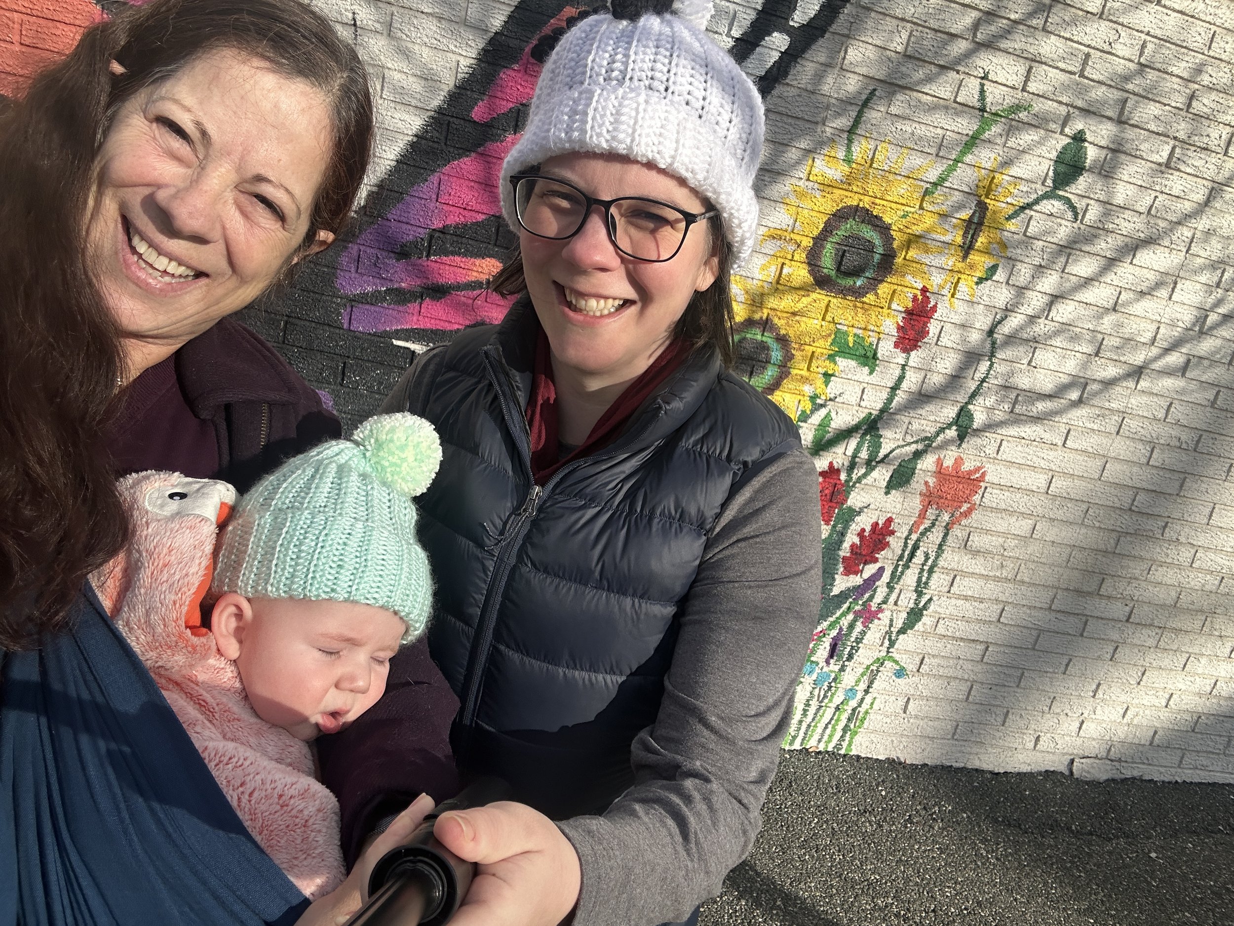 Linda Perry PMHNP-BC CNM holding her newborn grandson beside her daughter at an outdoor flower mural, reflecting her background in perinatal and reproductive mental health in NJ NY and MD