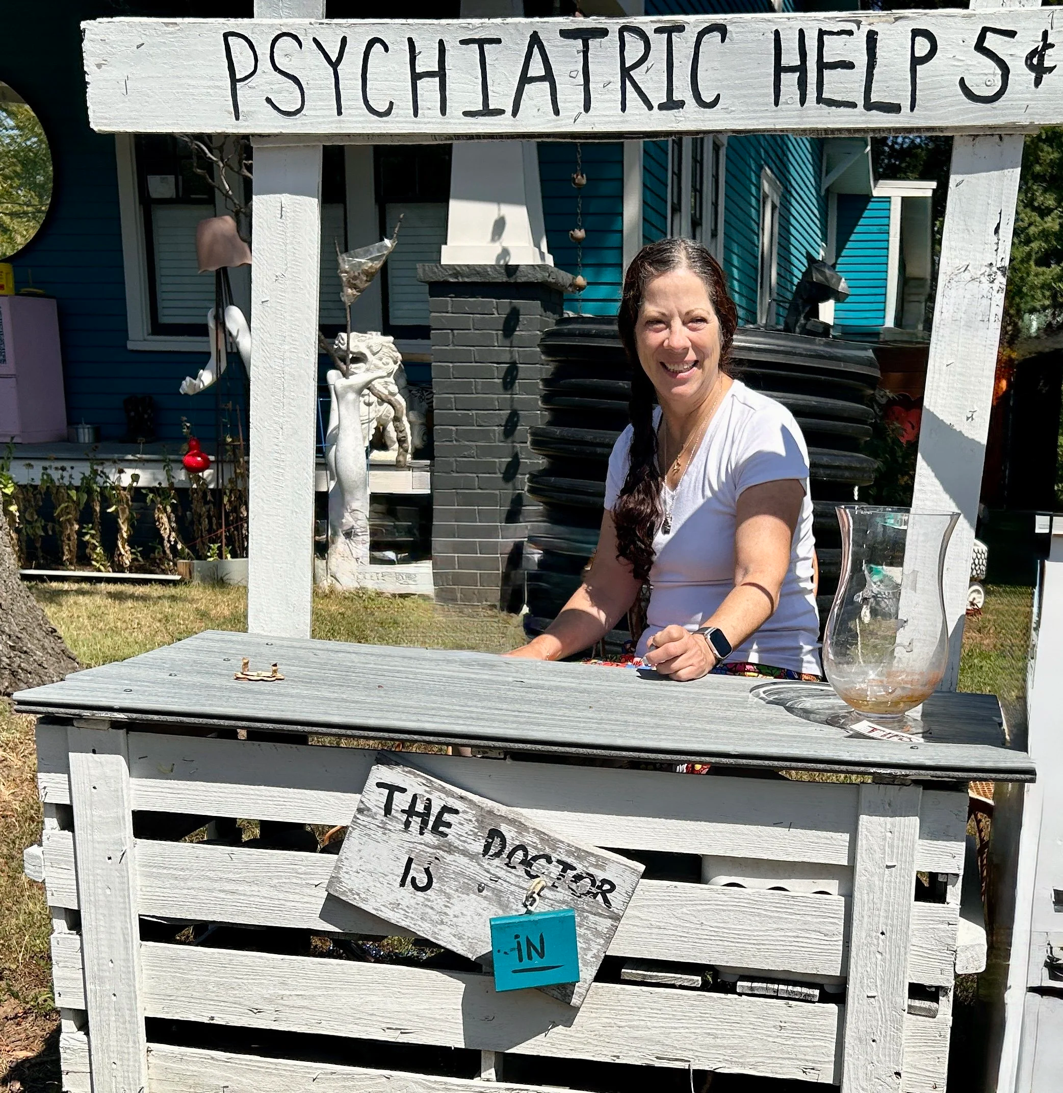 Linda Perry PMHNP-BC CNM smiling behind a hand-painted Psychiatric Help 5 cents booth with a The Doctor Is In sign, board-certified psychiatric nurse practitioner and certified nurse midwife serving NJ NY and MD
