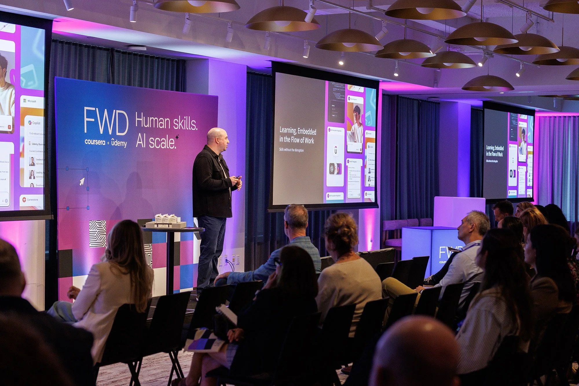 A man giving a presentation at a conference with a large screen behind him and audience members seated in front. The screen displays information about human skills and AI at scale, and the conference environment is decorated with purple and blue lighting.