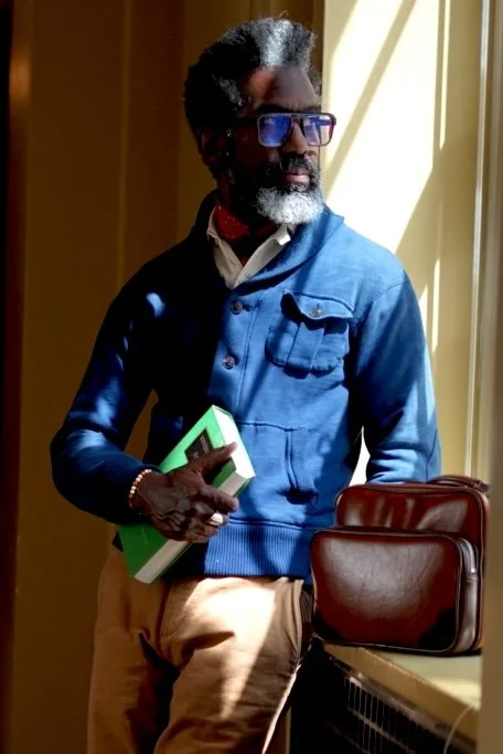 Man with glasses, gray beard, and gray hair, wearing a blue jacket, holding a green book, standing by a window with sunlight streaming in.