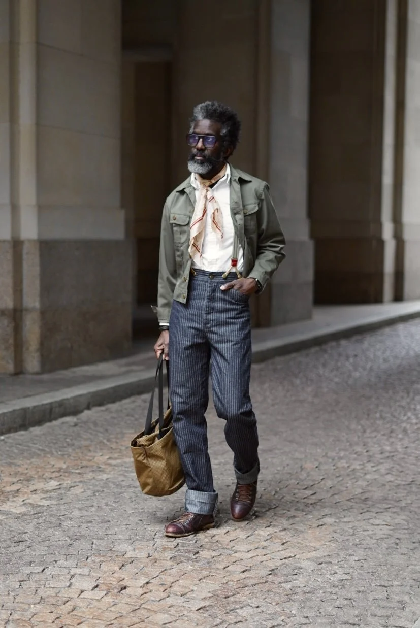 A stylish man walking on cobblestone sidewalk in an urban setting, holding a brown bag, wearing sunglasses, a green jacket over a light shirt with a scarf, pinstriped pants, and brown boots.