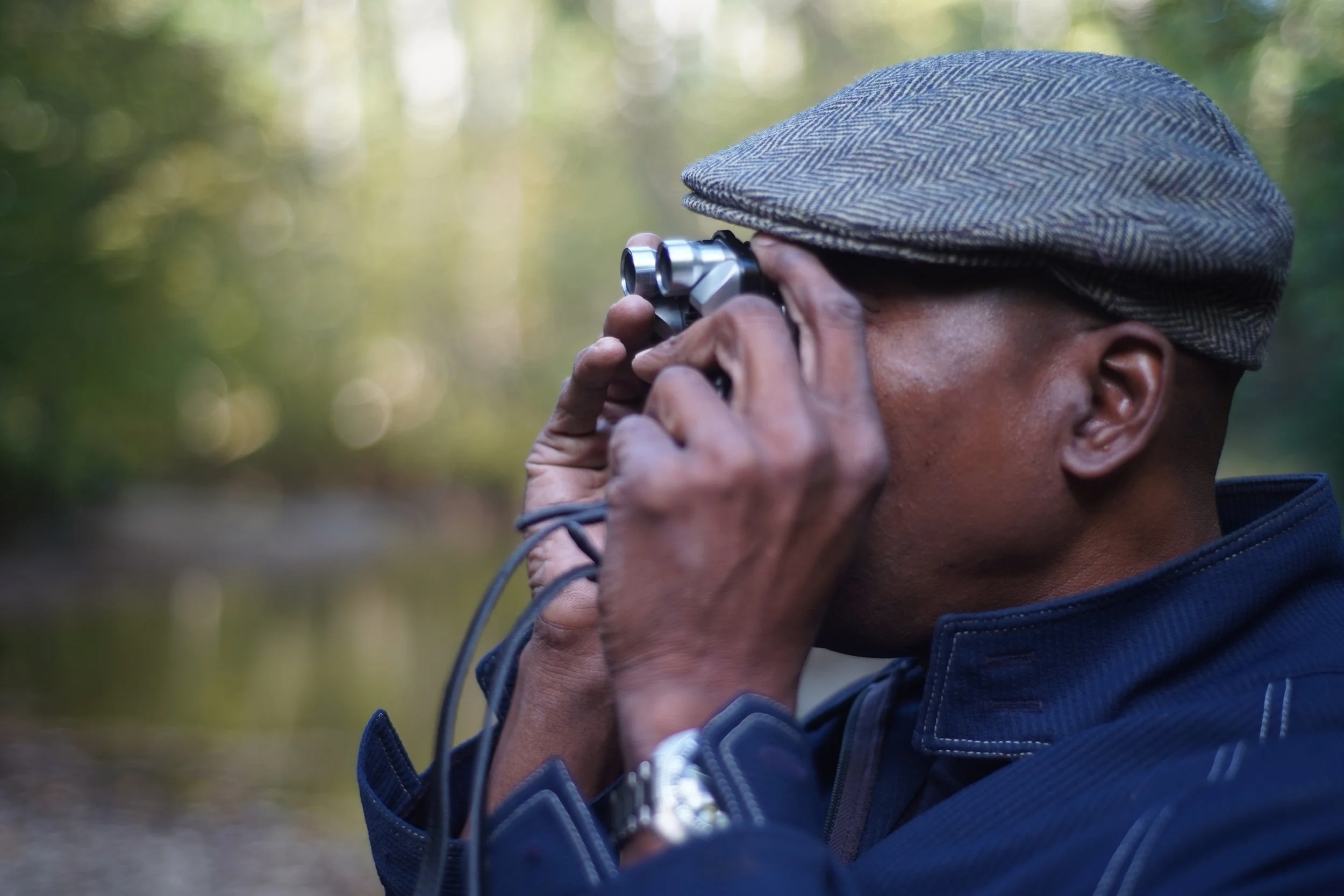 A person wearing a plaid cap using binoculars outdoors in a wooded area.