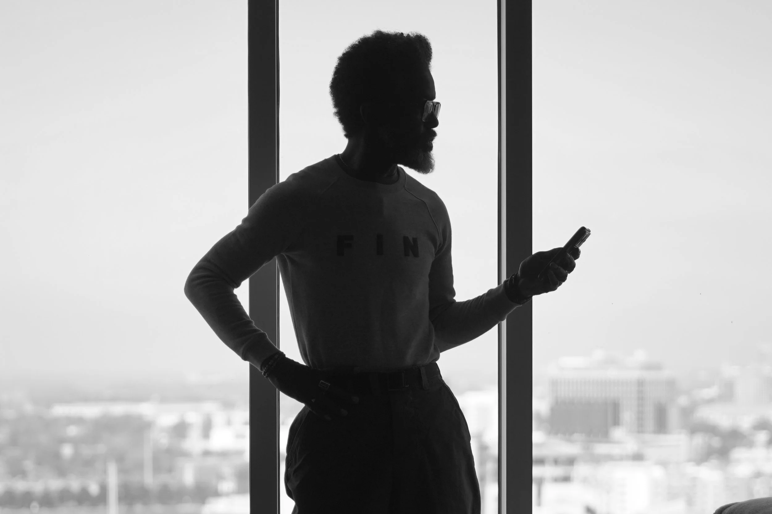 Silhouette of a man with glasses holding a smartphone in front of a large window with a city view.