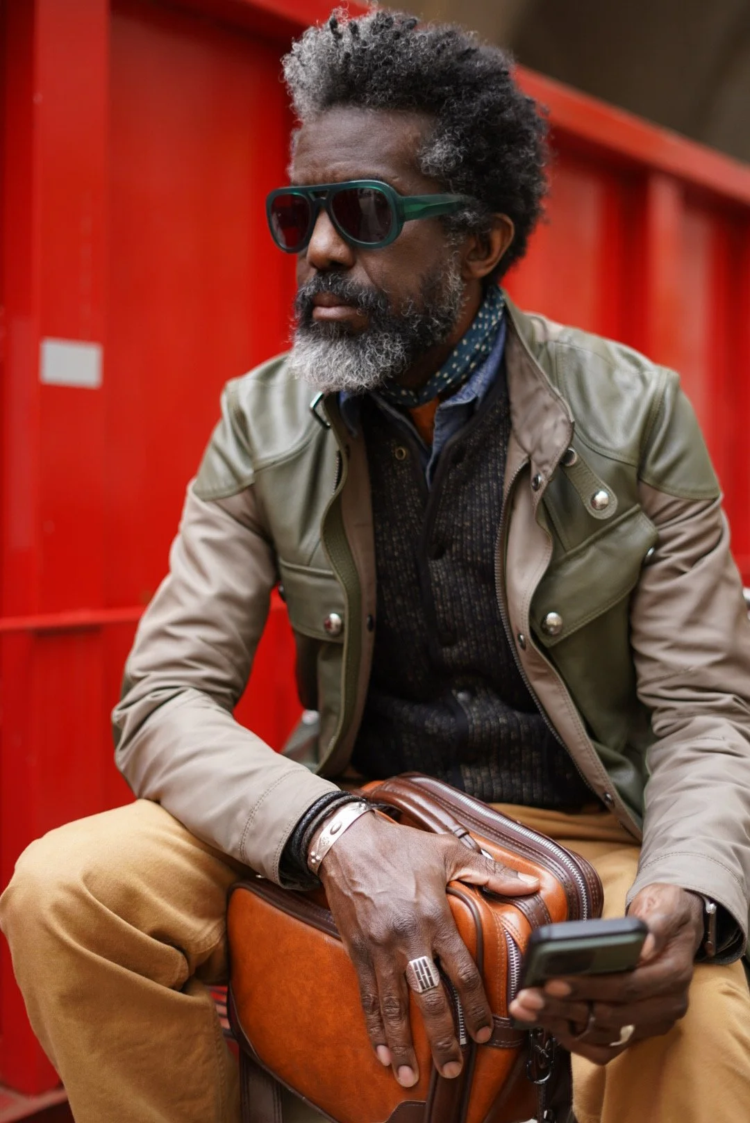 A middle-aged man with gray hair and beard, wearing dark sunglasses, a layered jacket, and tan pants, sits outdoors holding a smartphone and a brown leather bag, with a red background.