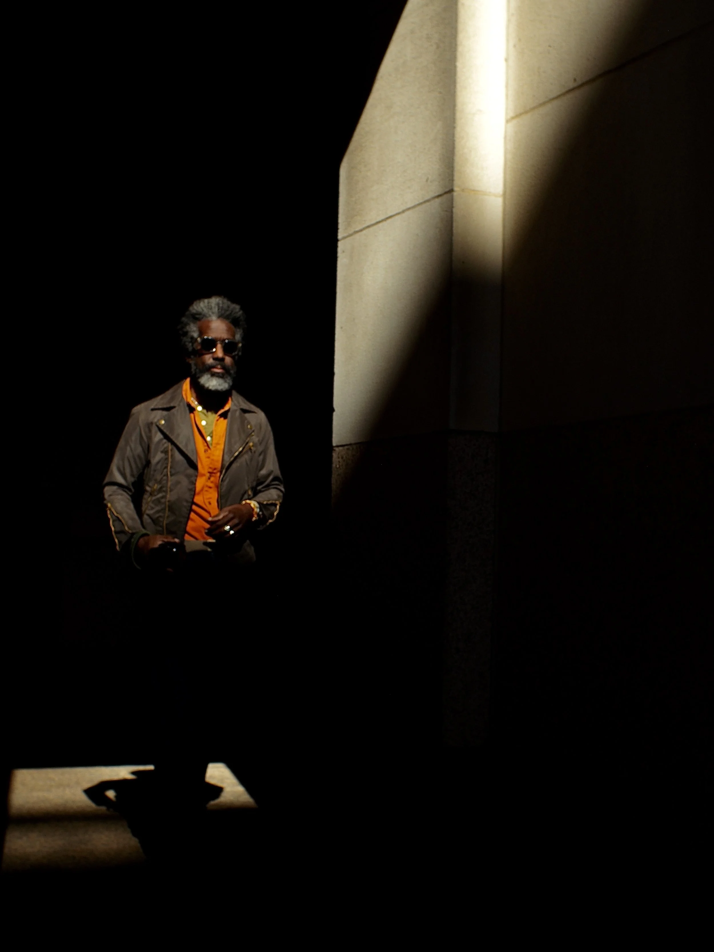 A man with gray hair and a beard, wearing sunglasses, an orange shirt, and a brown jacket, standing in a dark space with light shining on a stone wall behind him.