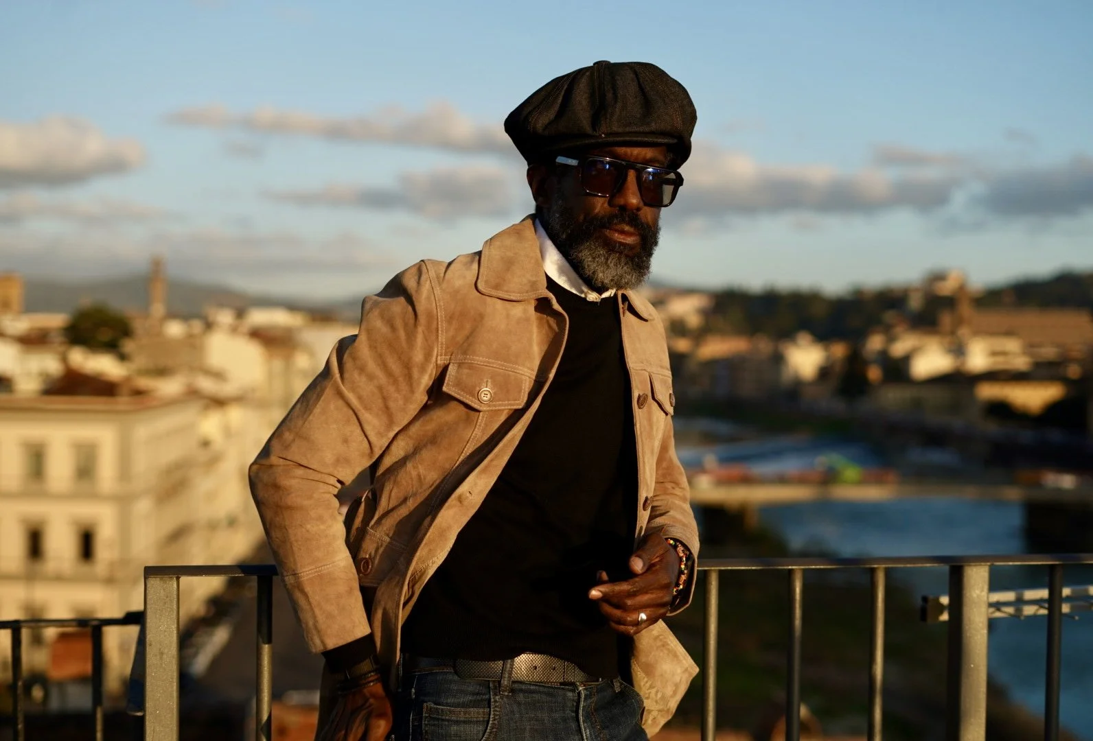 A man with a beard wearing a brown leather jacket, black shirt, hat, and sunglasses is leaning on a railing with a cityscape and a river in the background during sunset.