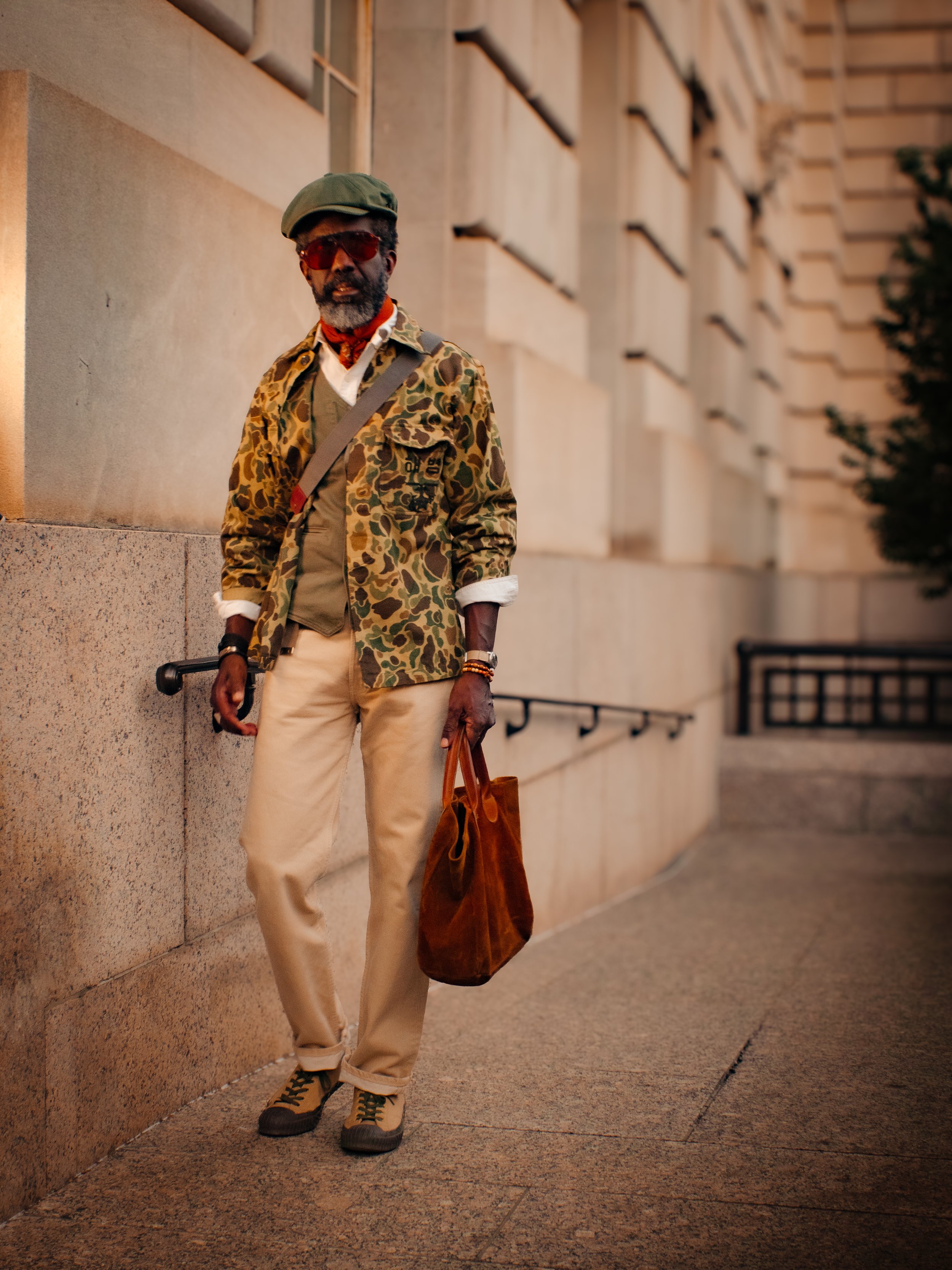 A man with gray hair and a beard standing outdoors at night, leaning against a beige stone building wall. He is wearing a green cap, red-tinted sunglasses, a leopard print jacket, beige pants, and brown shoes. He is holding a brown bag in one hand and has a shoulder strap across his chest.