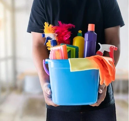 Person holding a blue cleaning bucket filled with various cleaning supplies including spray bottles, sponges, cloths, and cleaning brushes.