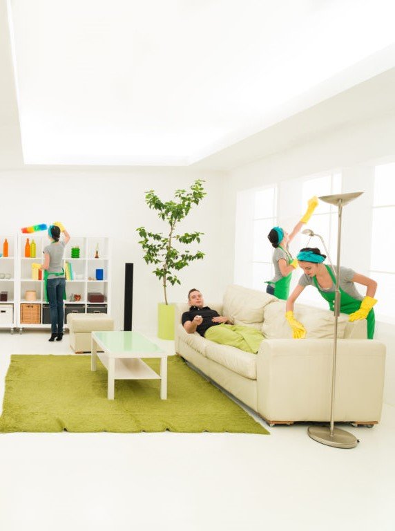 Two women wearing cleaning gloves and headphones cleaning the living room while a man lies on the sofa, relaxing.