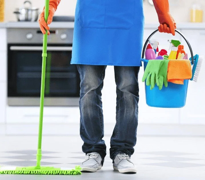 Person in a blue apron and jeans holding a cleaning bucket with cleaning supplies and wearing orange gloves, standing on a kitchen floor.