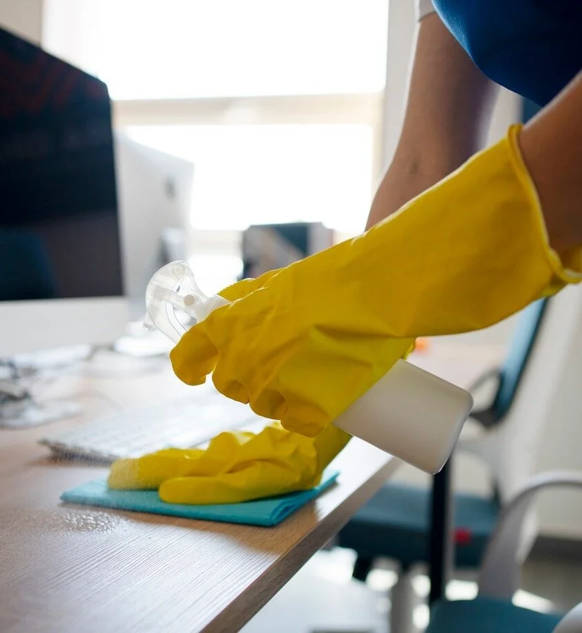Person wearing yellow rubber gloves cleaning a desk with a cloth and spray bottle in an office setting.