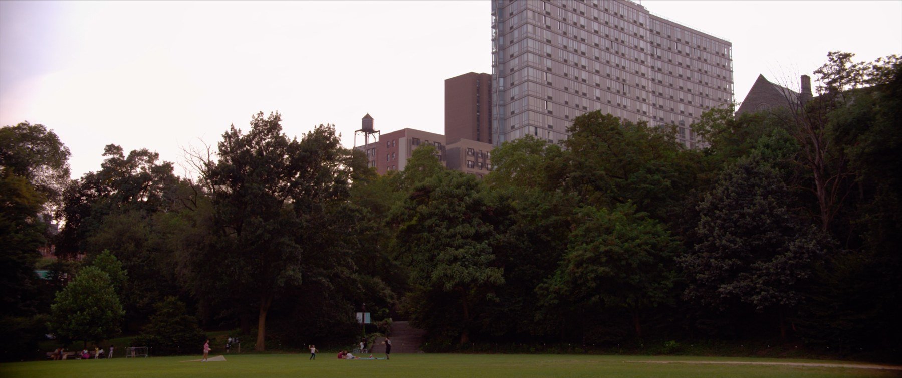 Quietly short film still — Kai Wen Hu - A park with children playing on the grass, surrounded by trees, with tall buildings in the background under a partly cloudy sky.