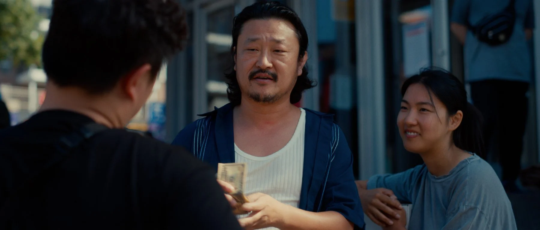 Pedigree short film still — Kai Wen Hu - Samuel and Squid engaged in a cash exchange on a city street, smiling.