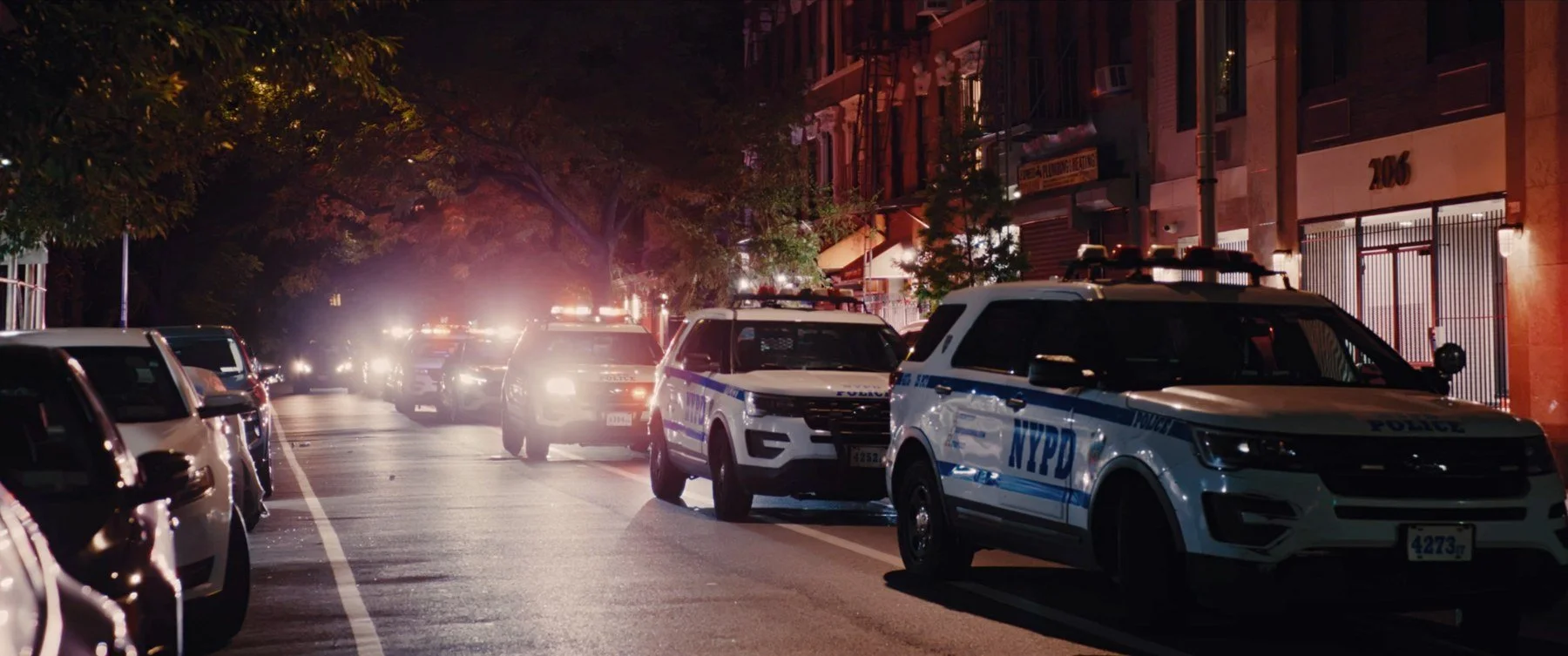 Quietly short film still — Kai Wen Hu - Nighttime city street with multiple police cars parked along the curb, illuminated by streetlights and vehicle headlights, with buildings and trees visible in the background.