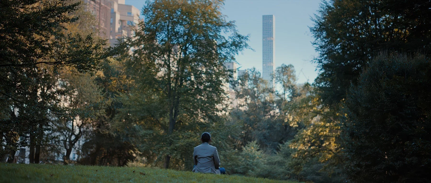 Pedigree short film still — Kai Wen Hu - Samuel sits in a park overlooked by a tall, narrow skyscraper filled with luxury condos.