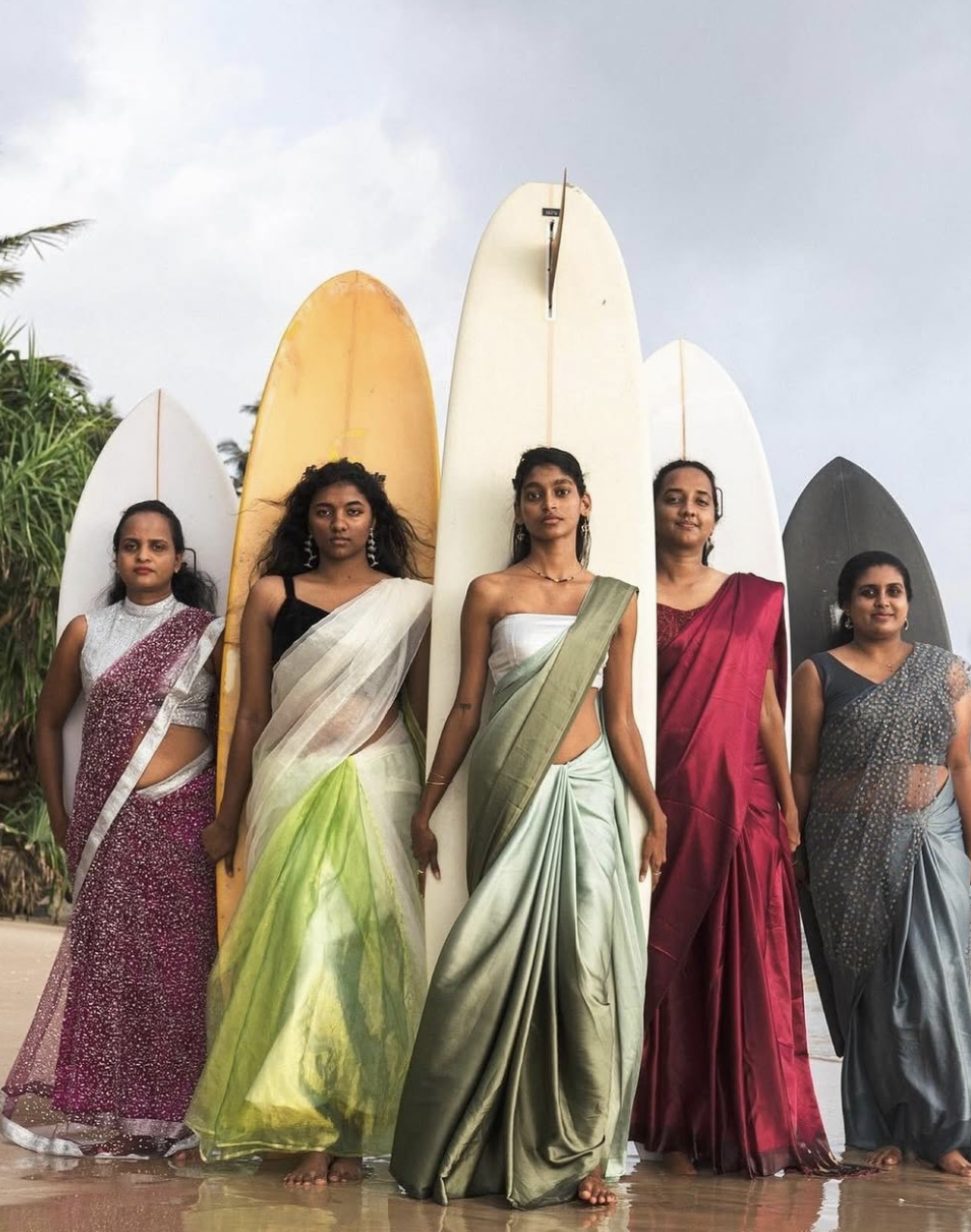 5 Sri Lankan women wearing saris, standing in front of their surf boards on the beach