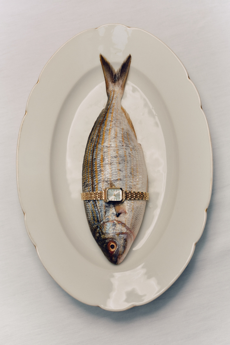 A whole uncooked fish on a white platter, with a gold watch around the fish's belly