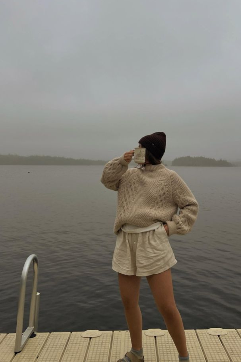 Woman standing at the edge of a dock, wearing a cream knit fisherman sweater, knit beanie, linen shorts, and socks with sandals, holding a mug up to her face