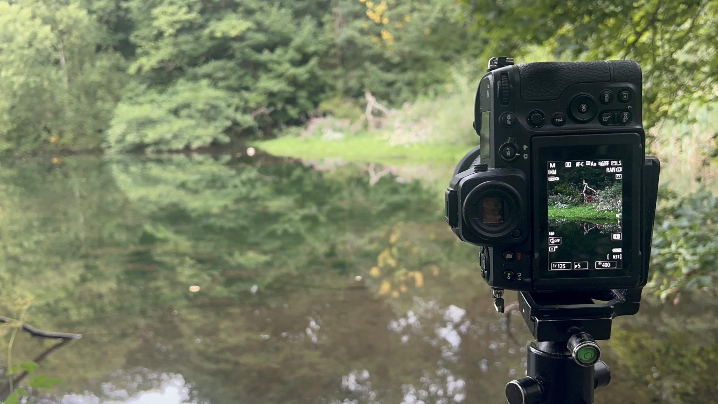 A digital camera mounted on a tripod taking a picture of a green landscape with trees and a body of water.