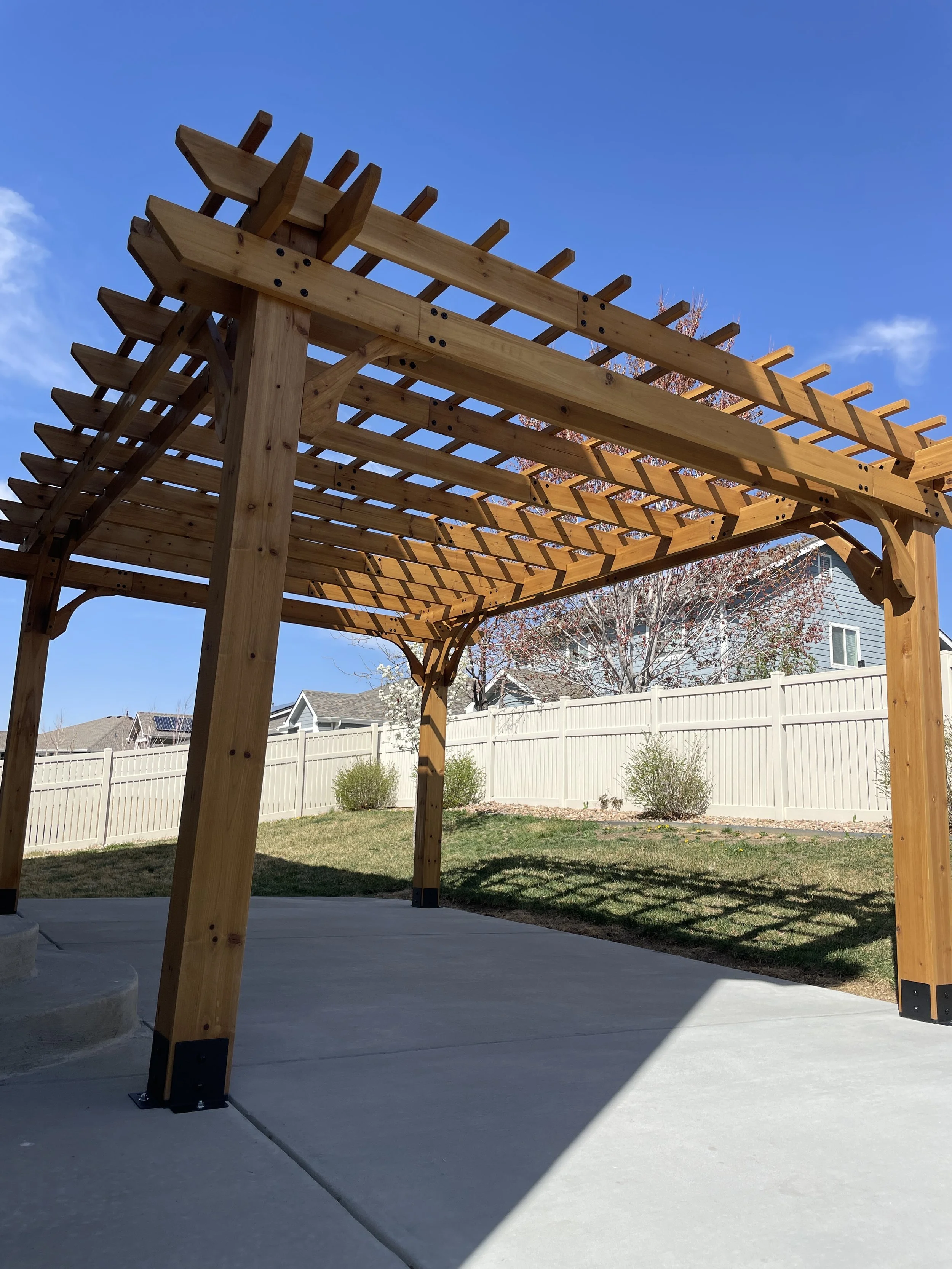 A wooden pergola with a lattice roof over a concrete patio in a backyard, with a white privacy fence and trees in the background.