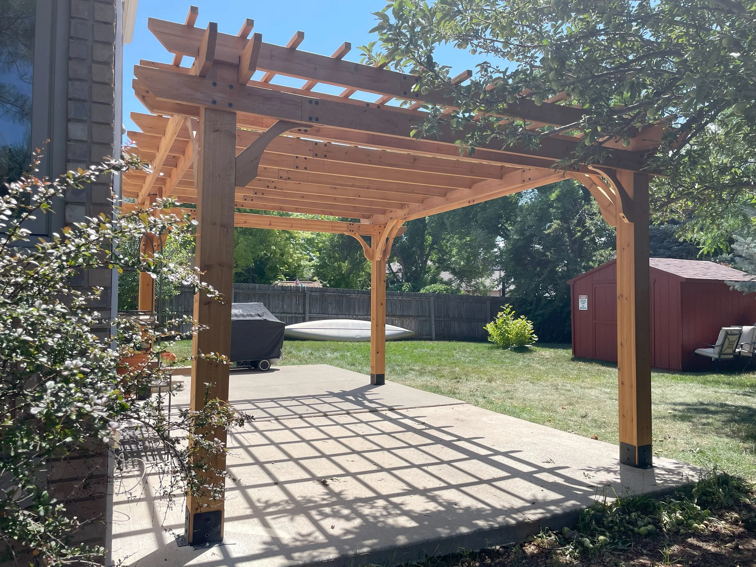 Backyard patio with a wooden pergola and concrete floor, surrounded by a wooden fence, trees, and garden shed.
