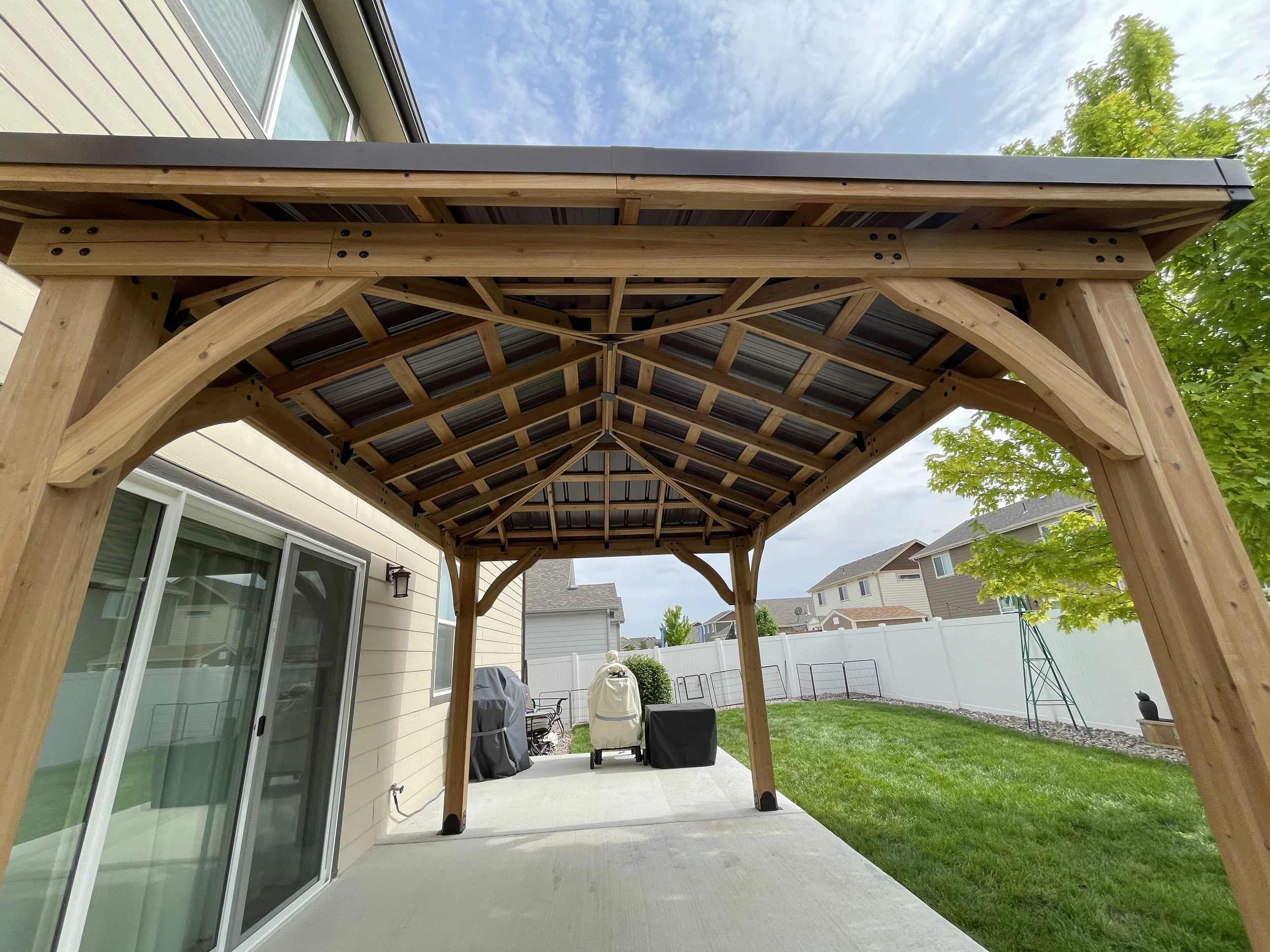 Wooden patio cover with a metal roof over a concrete patio in a backyard, with houses and trees in the background.