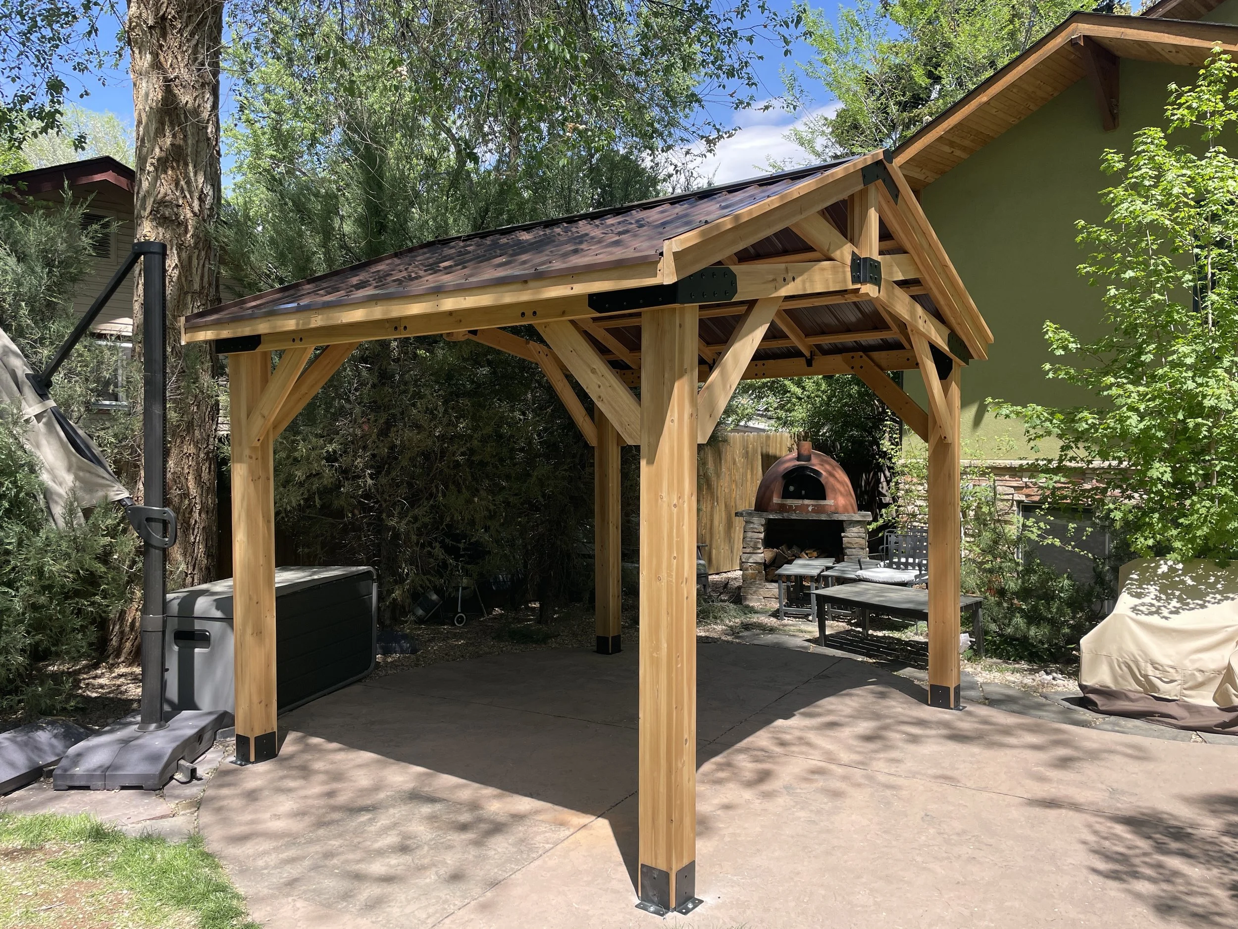 A wooden pergola with a brown metal roof in a backyard, with a brick pizza oven, outdoor seating, a covered grill, and trees surrounding the area.