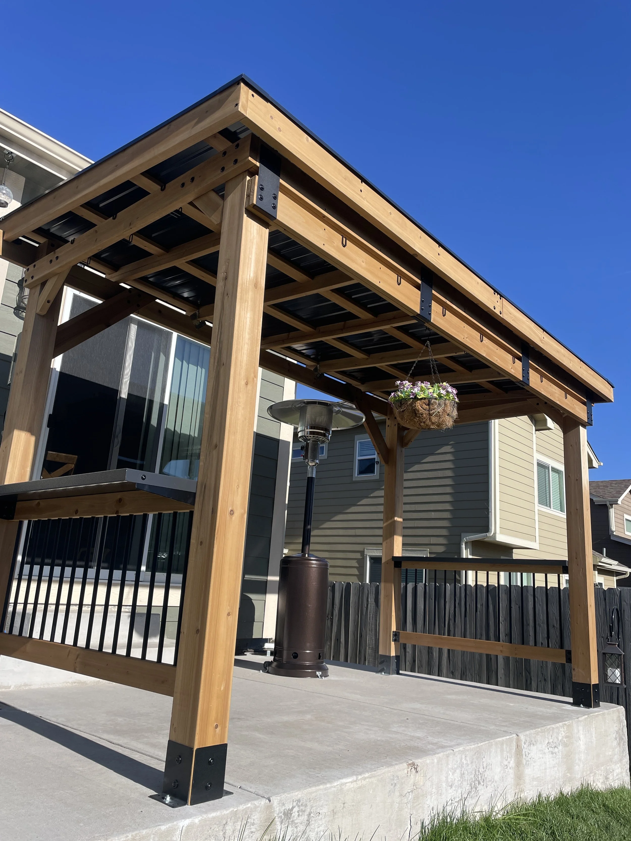 Wooden backyard deck with a metal railing, hanging flower basket, outdoor patio heater, and black fence in the background under a clear blue sky.