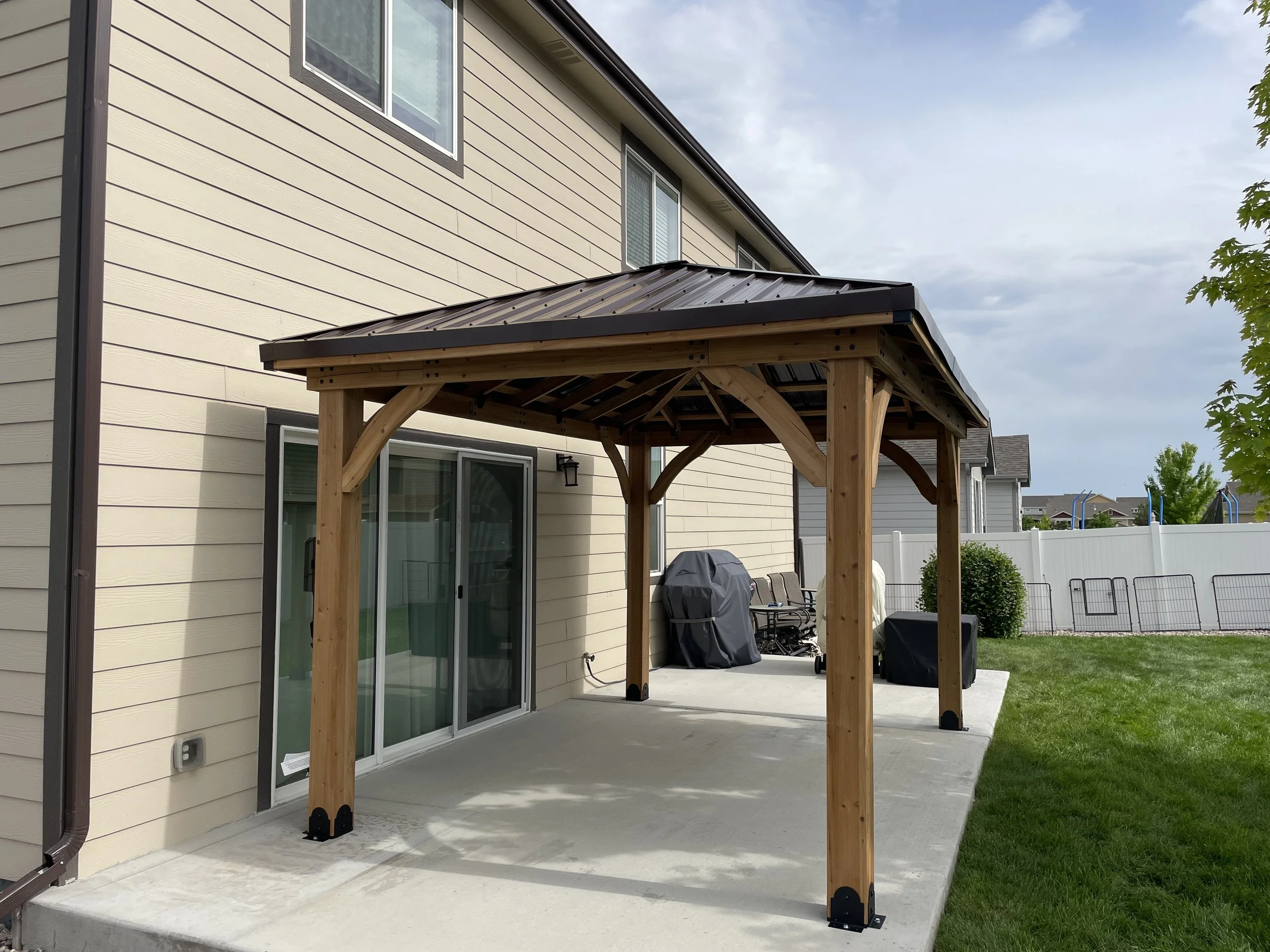 Backyard patio with a wooden pergola, sliding glass door, barbecue grill, outdoor chairs, and a grassy yard with a white fence.