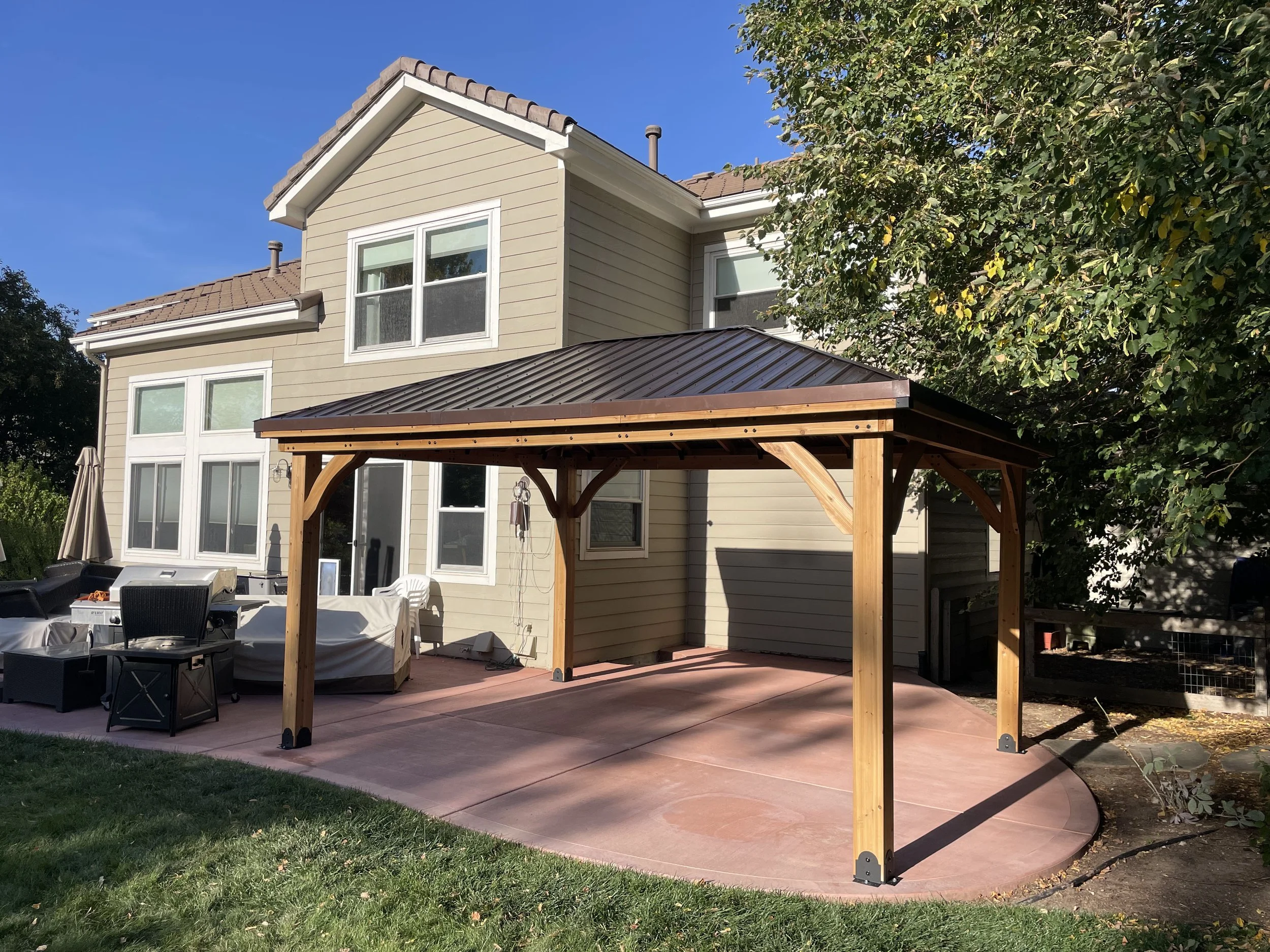 A backyard patio with a wooden pergola attached to a beige two-story house, with outdoor furniture and a large tree providing partial shade.