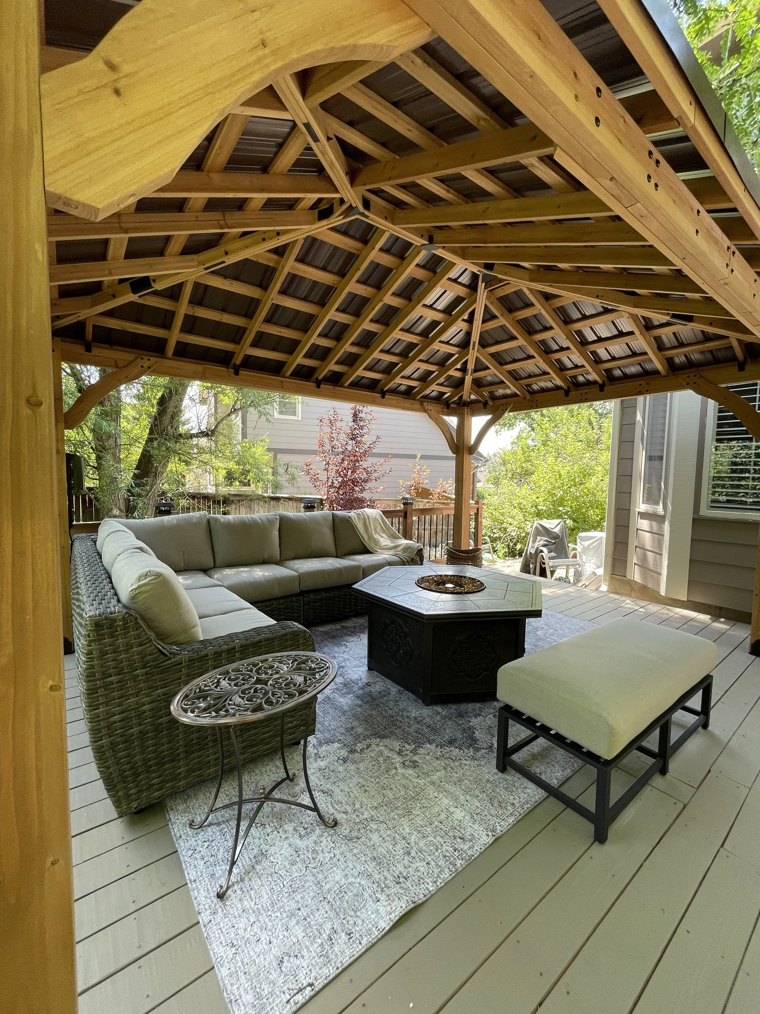 Covered outdoor patio with a large beige sectional sofa, a black coffee table, an off-white cushioned bench, and a small metal side table on a light-colored wooden deck, with trees and a neighboring house visible in the background.