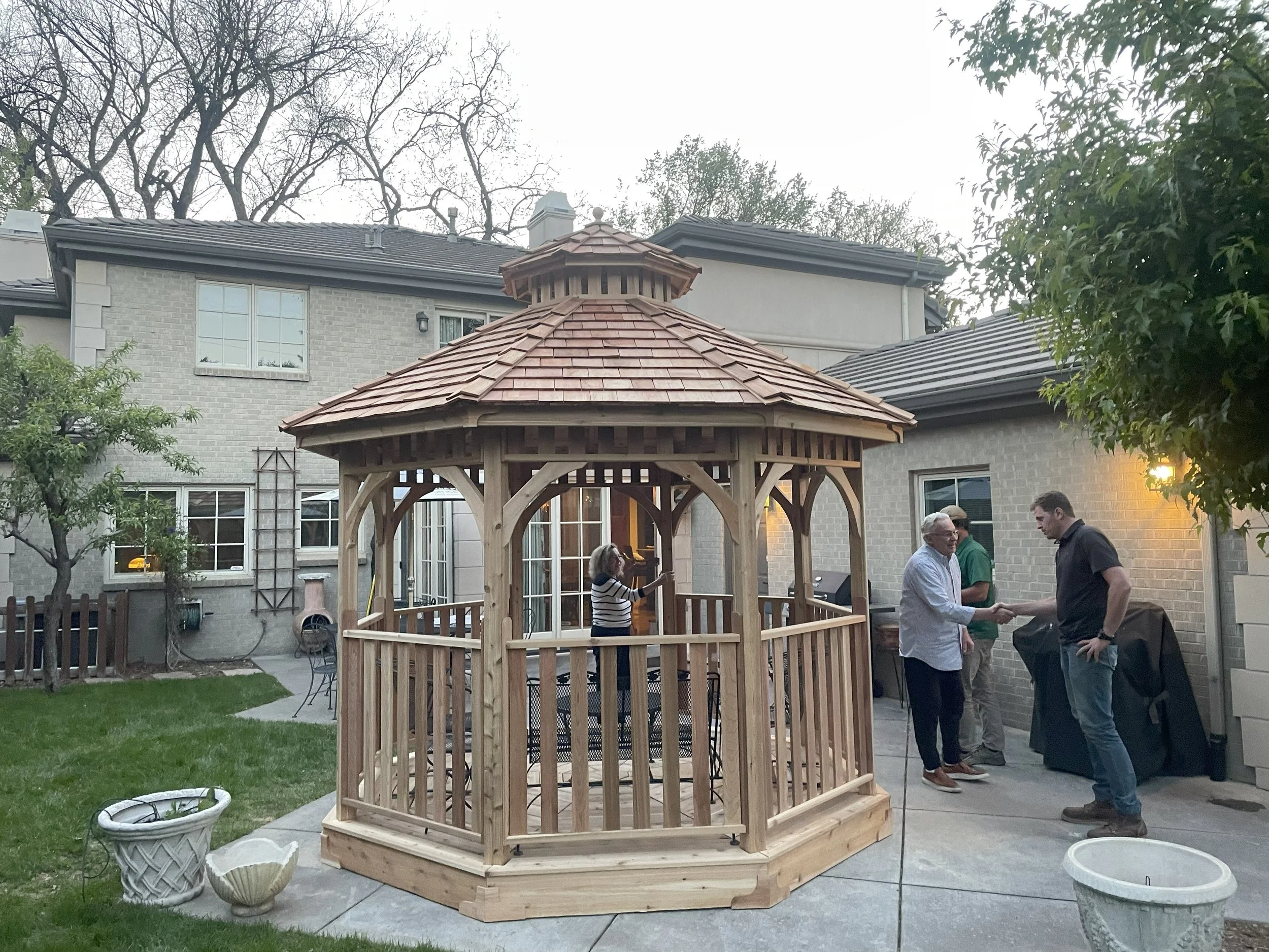 People in a backyard around a newly built wooden gazebo with a shingled roof, shaking hands and talking. There are potted plants and outdoor furniture nearby.