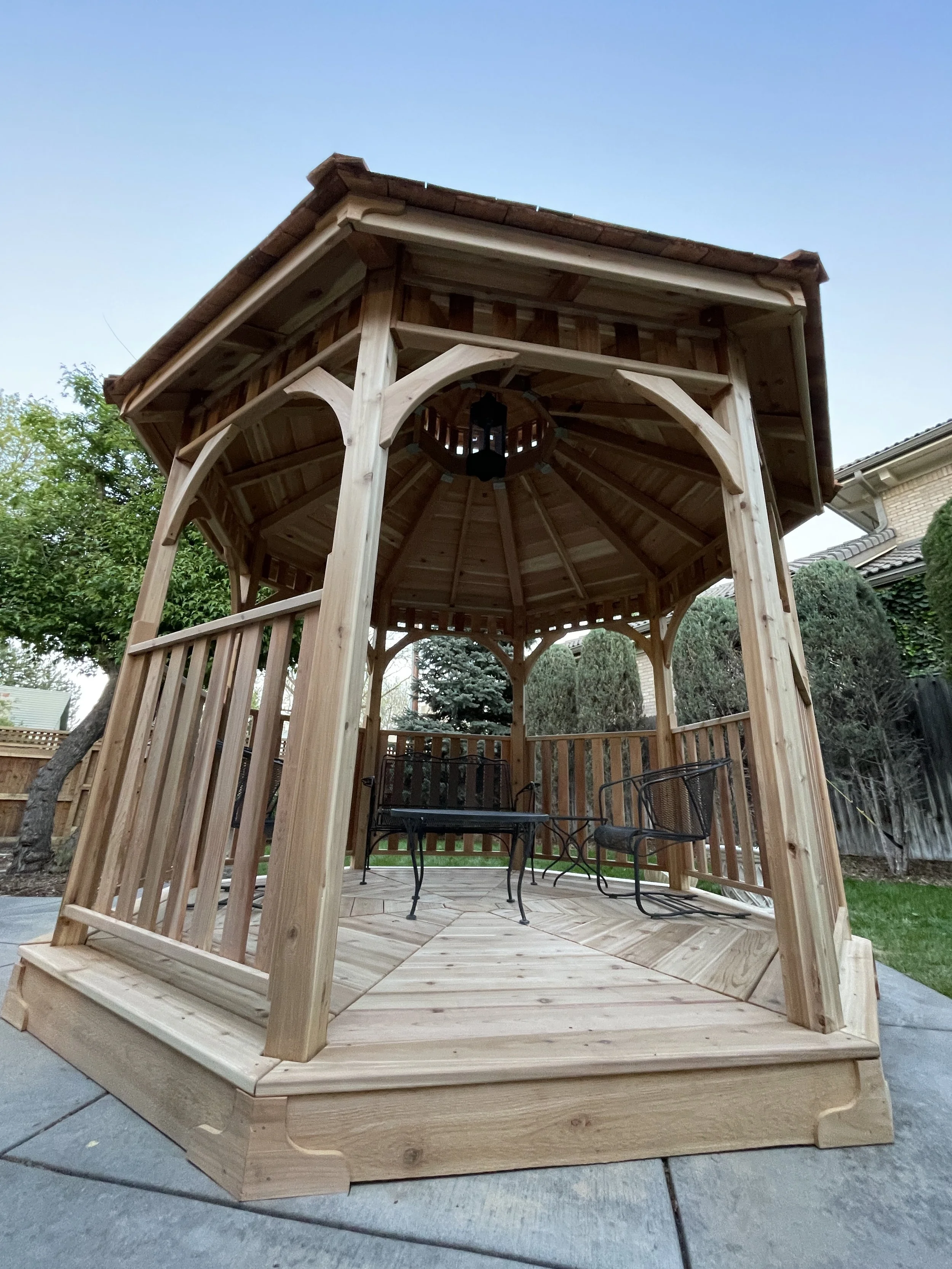A wooden gazebo with a hexagonal shape, elevated slightly above the ground, with a sitting area and a small table and chairs inside, located in a backyard with trees, bushes, and a house nearby.