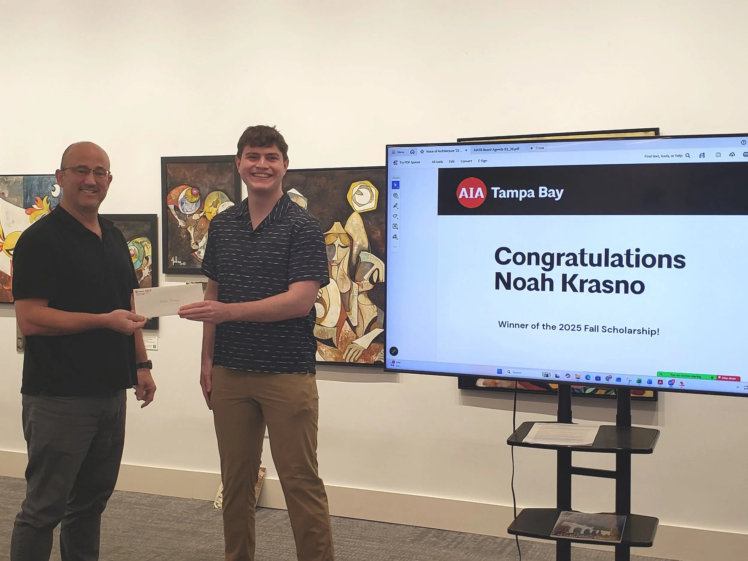 Scott Holmes, AIA President, handing a scholarship check to Noah Krasno, 2025 Fall AIA Scholarship Recipient. The monitor displays a message congratulating Noah Krasno as the winner of the 2025 Fall Scholarship from AIA Tampa Bay.