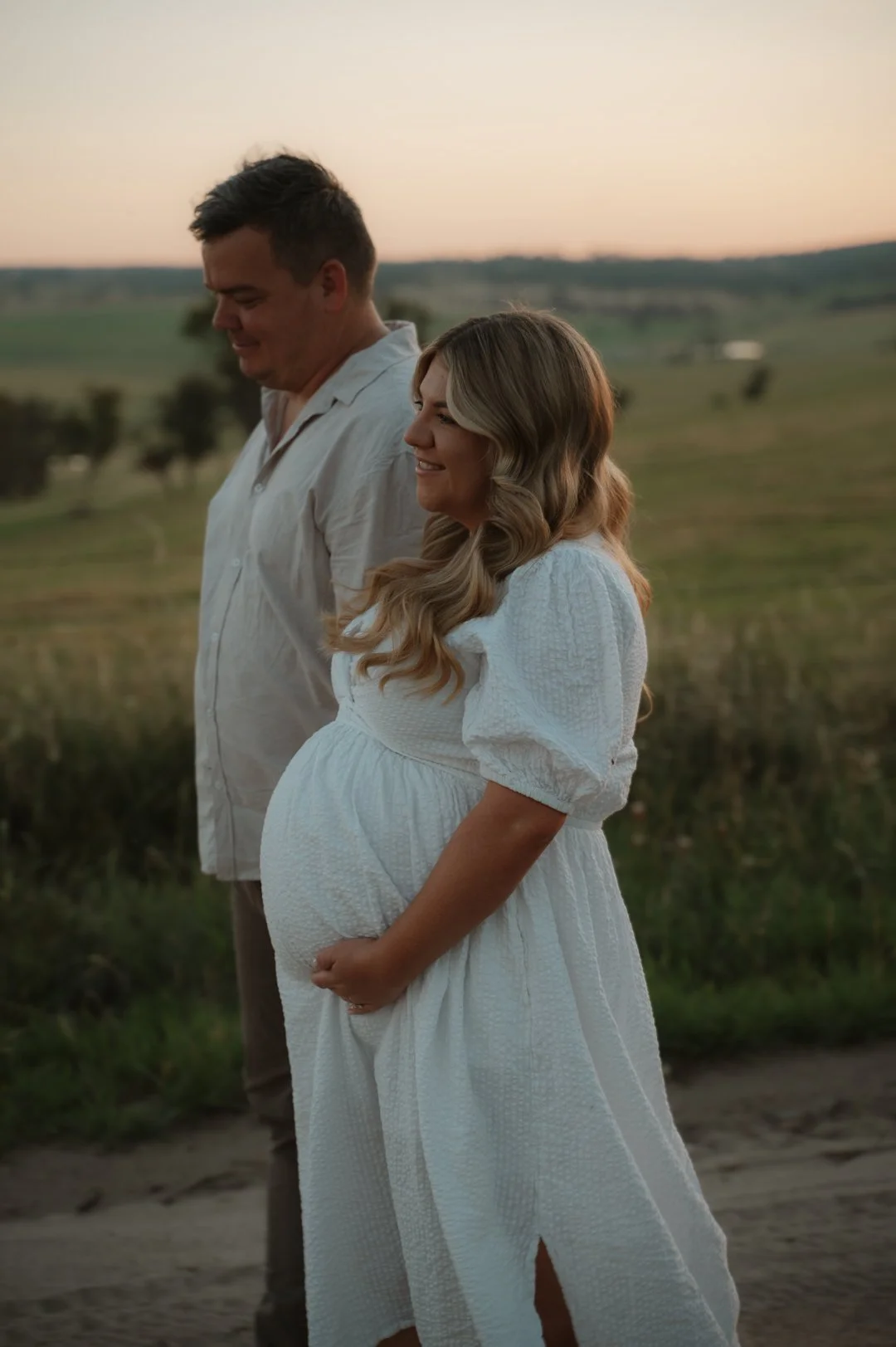 A pregnant woman in a white dress stands beside a man in a light-colored shirt in a grassy outdoor setting at sunset.