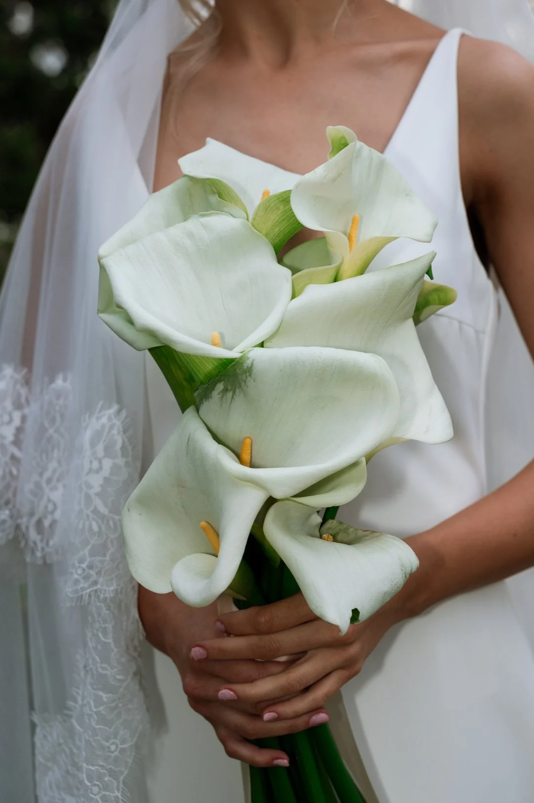 Close-up of a bride holding a bouquet of white calla lilies in her hands, wearing a white dress with lace details on the sleeve.