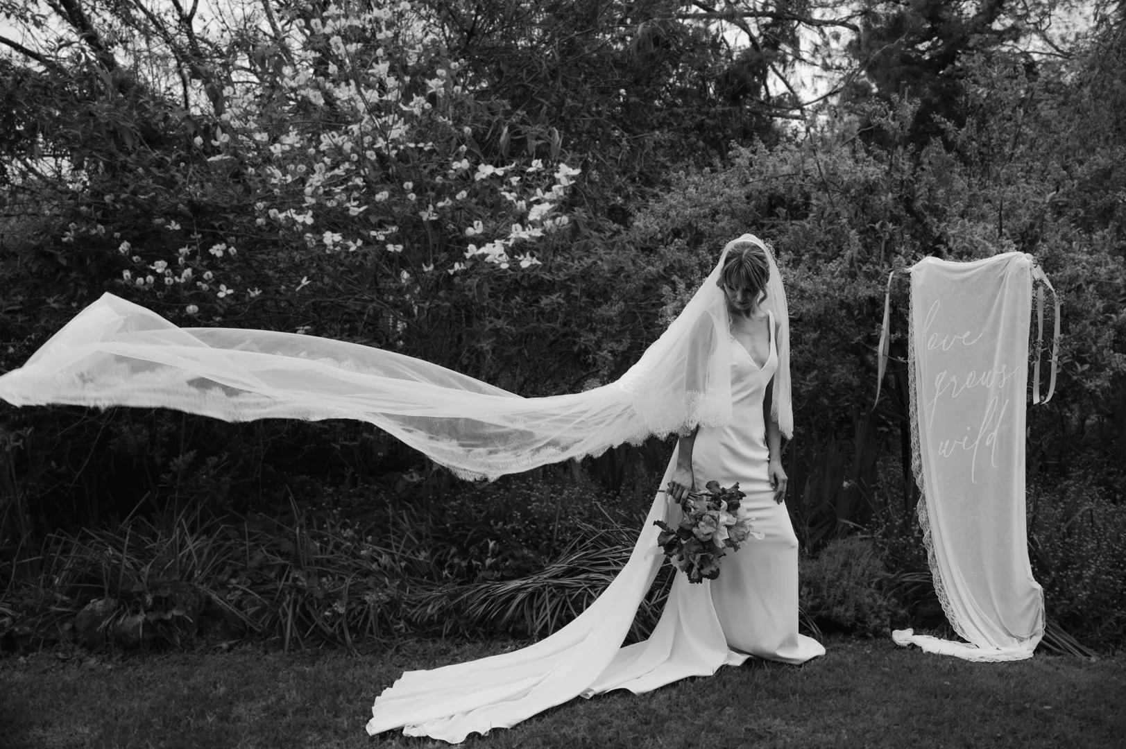 A bride in a wedding dress with a long veil standing outdoors, holding a bouquet, near a sign that says 'love grows wild'.
