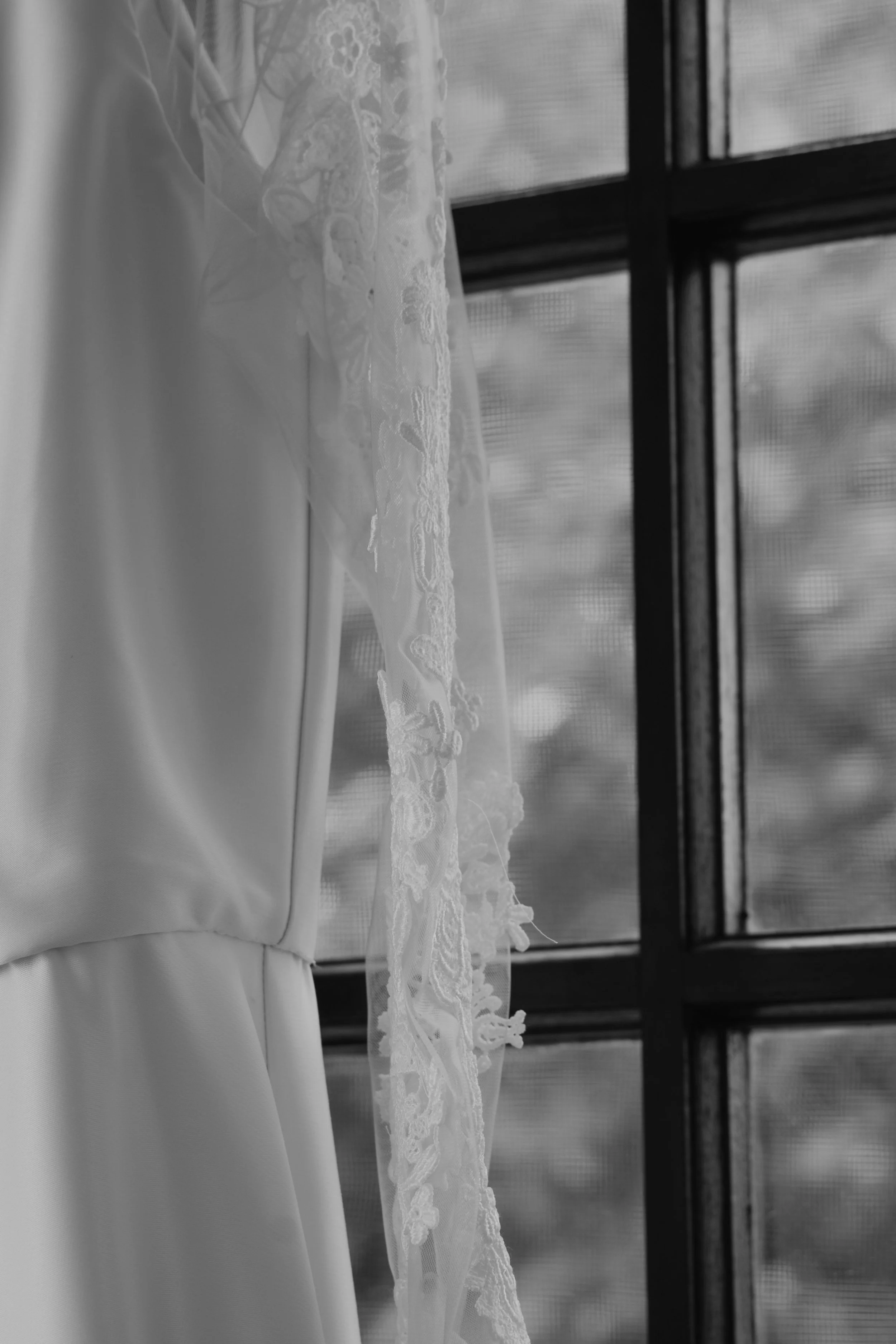 A close-up black and white photo of a wedding dress hanging by a window with lace details on the veil.
